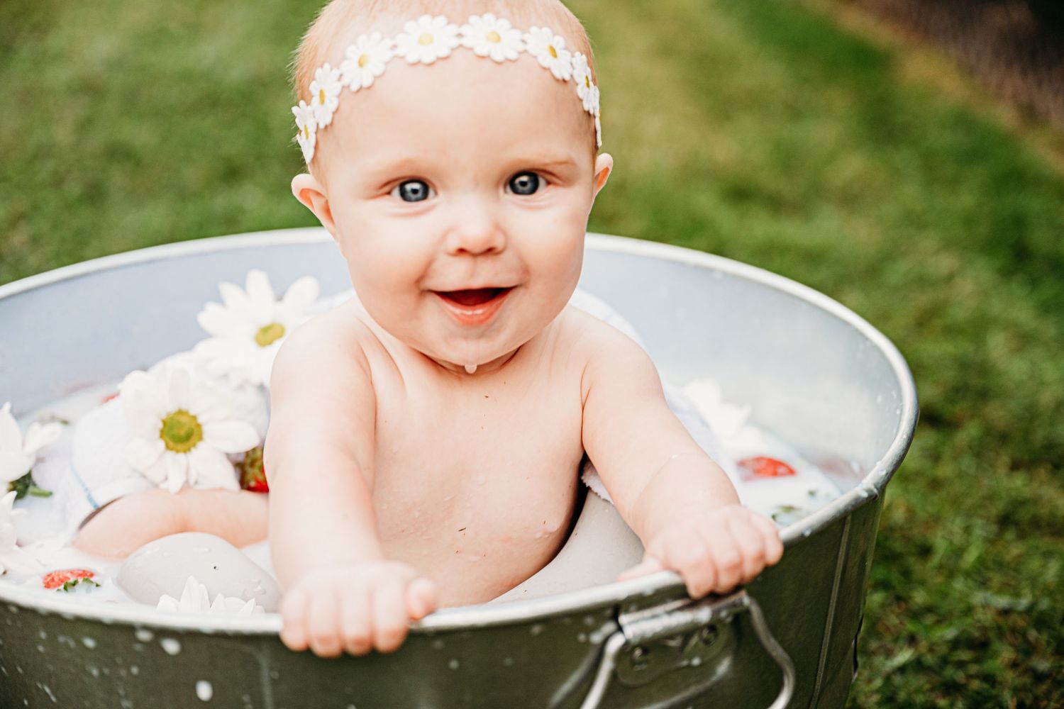 A baby smiling during an outdoor milk bath photography session with scattered daisies and flowers.