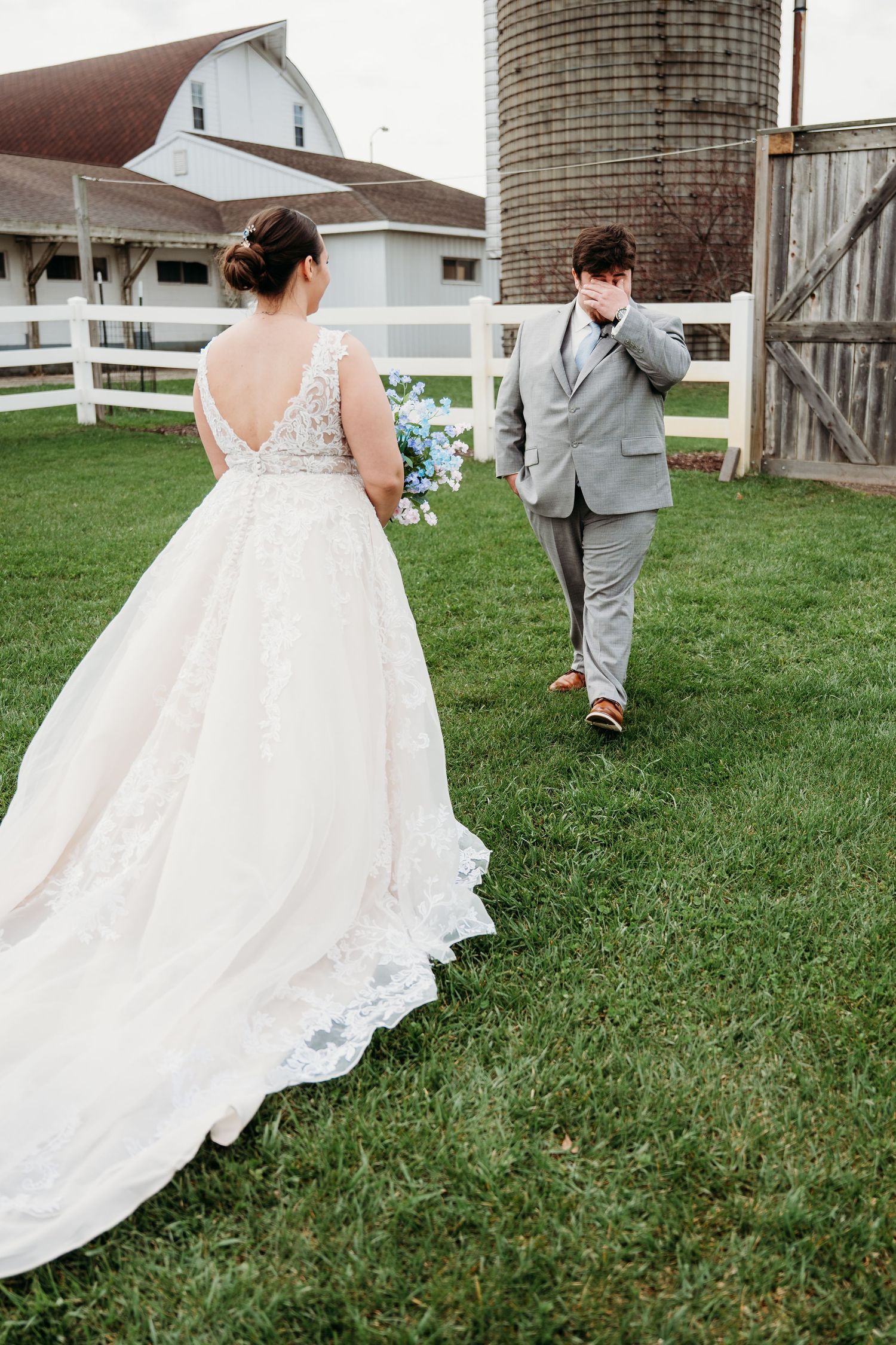 A couple in wedding attire walks through green grass near a rustic barn with a silo in the background.