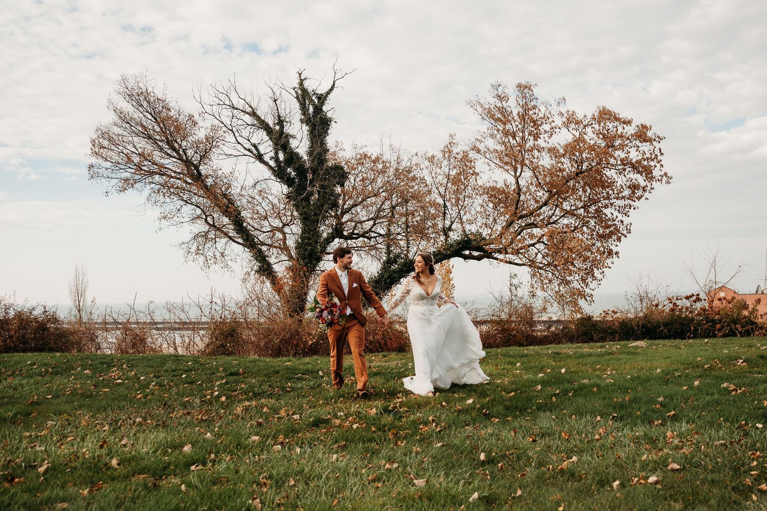 A couple stands together in a grassy field with autumn trees in the background during a romantic outdoor photo session.