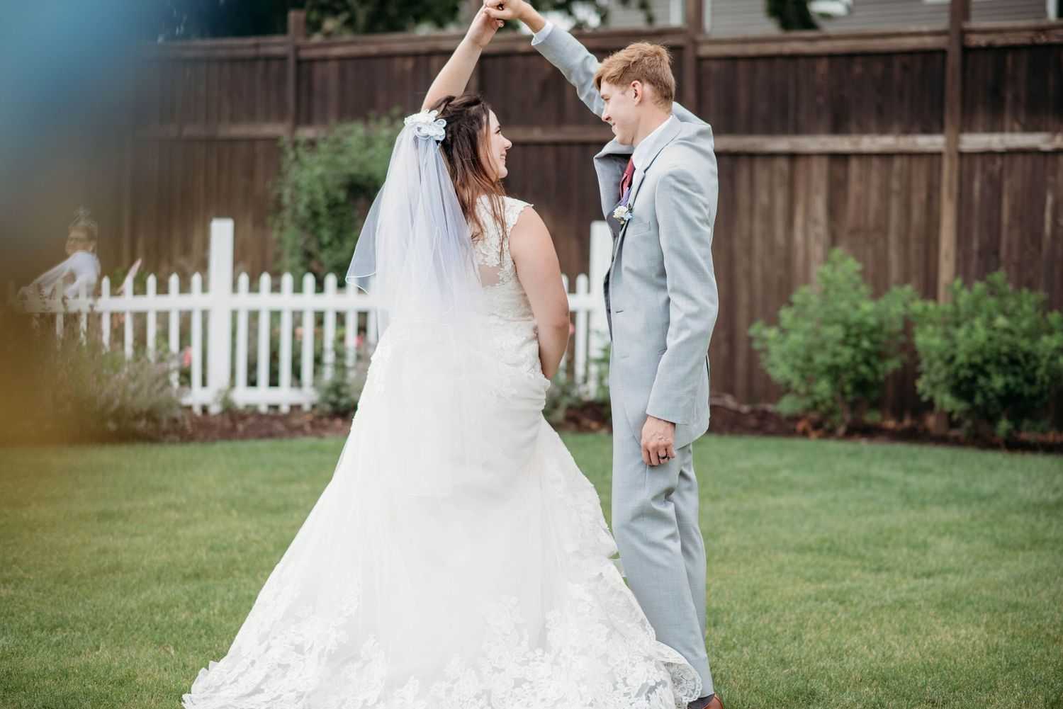 A romantic wedding moment captured in a backyard garden setting with a white picket fence and lush greenery backdrop.
