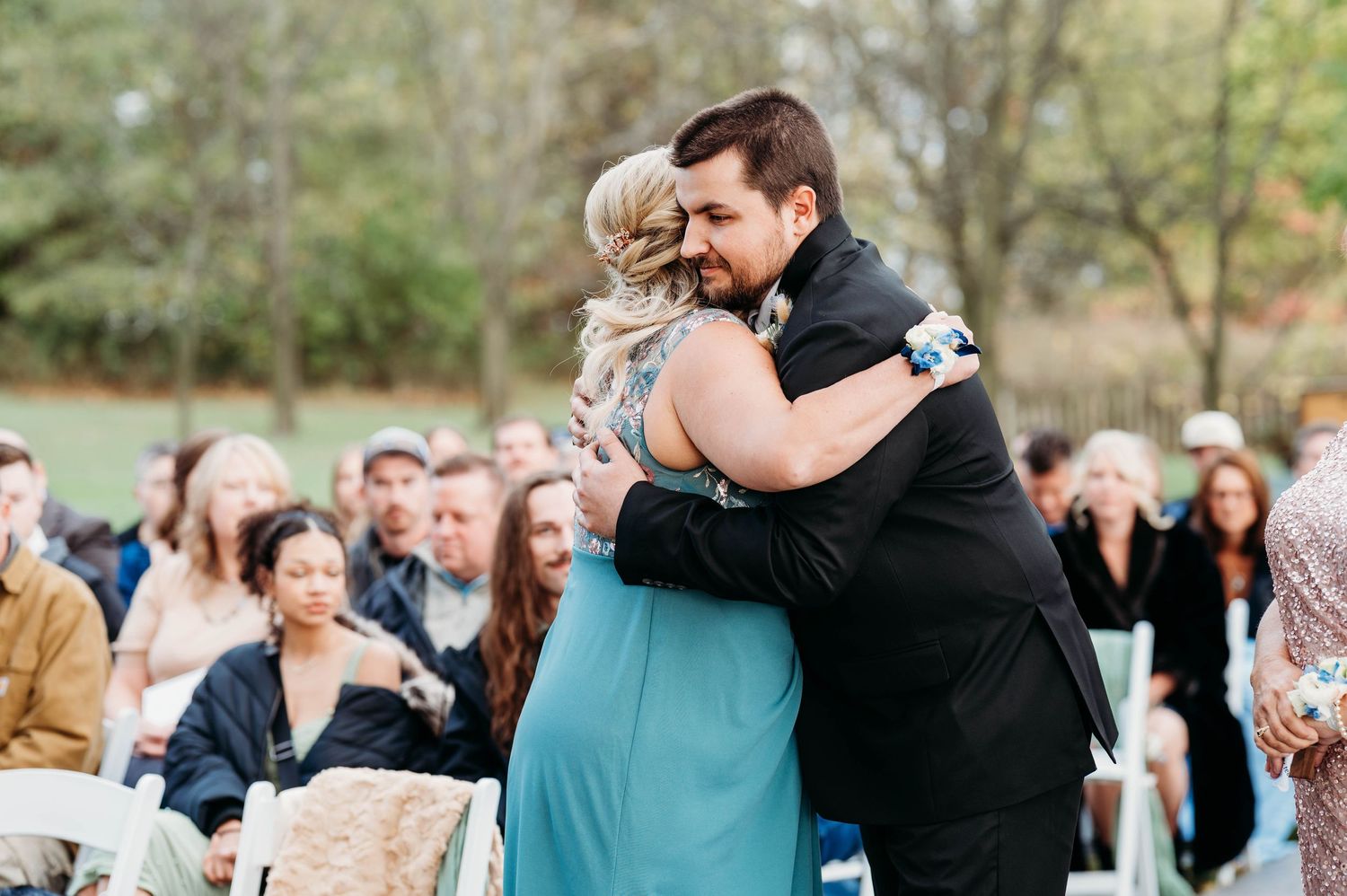 Groom in black tuxedo shares emotional hug with woman in teal dress during outdoor wedding ceremony.