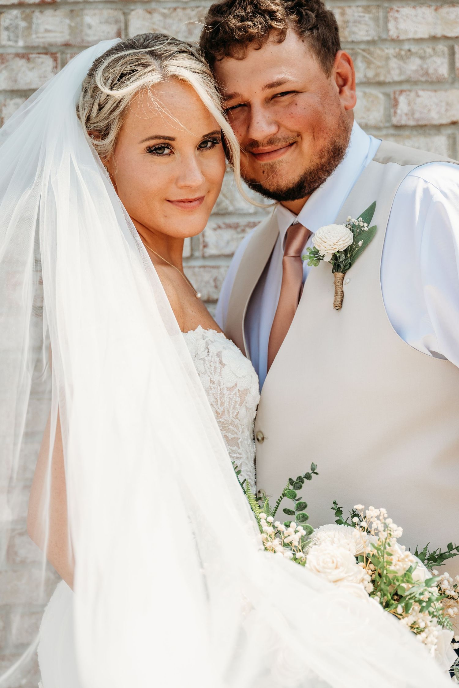 Bride and groom posing together on wedding day, bride wearing lace dress with veil holding white floral bouquet.