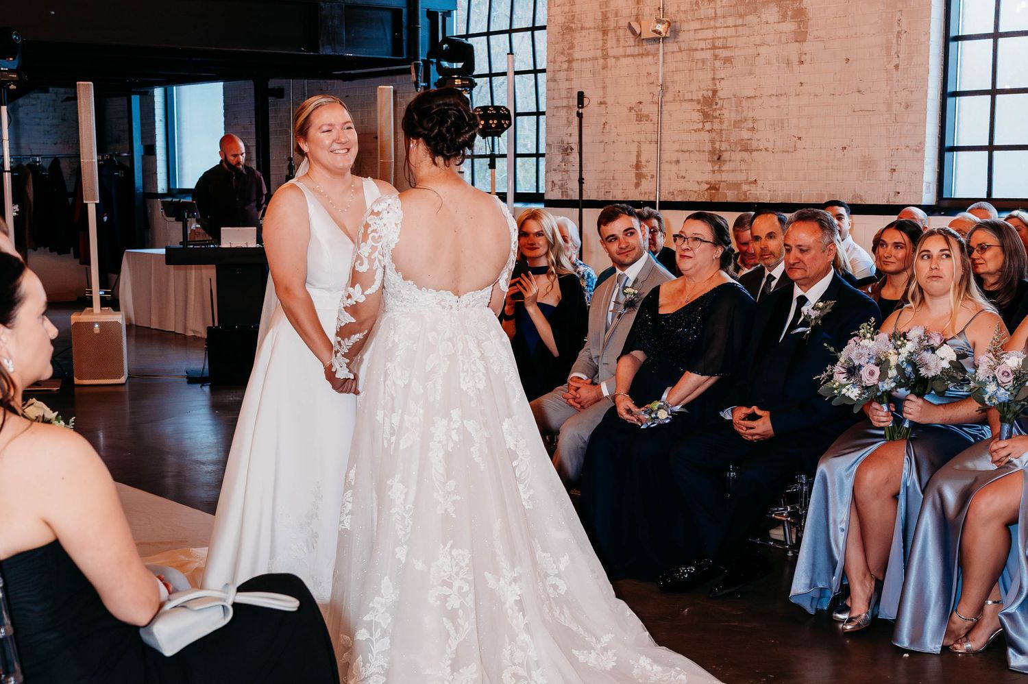 Two brides in white gowns exchange vows at an indoor wedding ceremony with guests seated nearby.