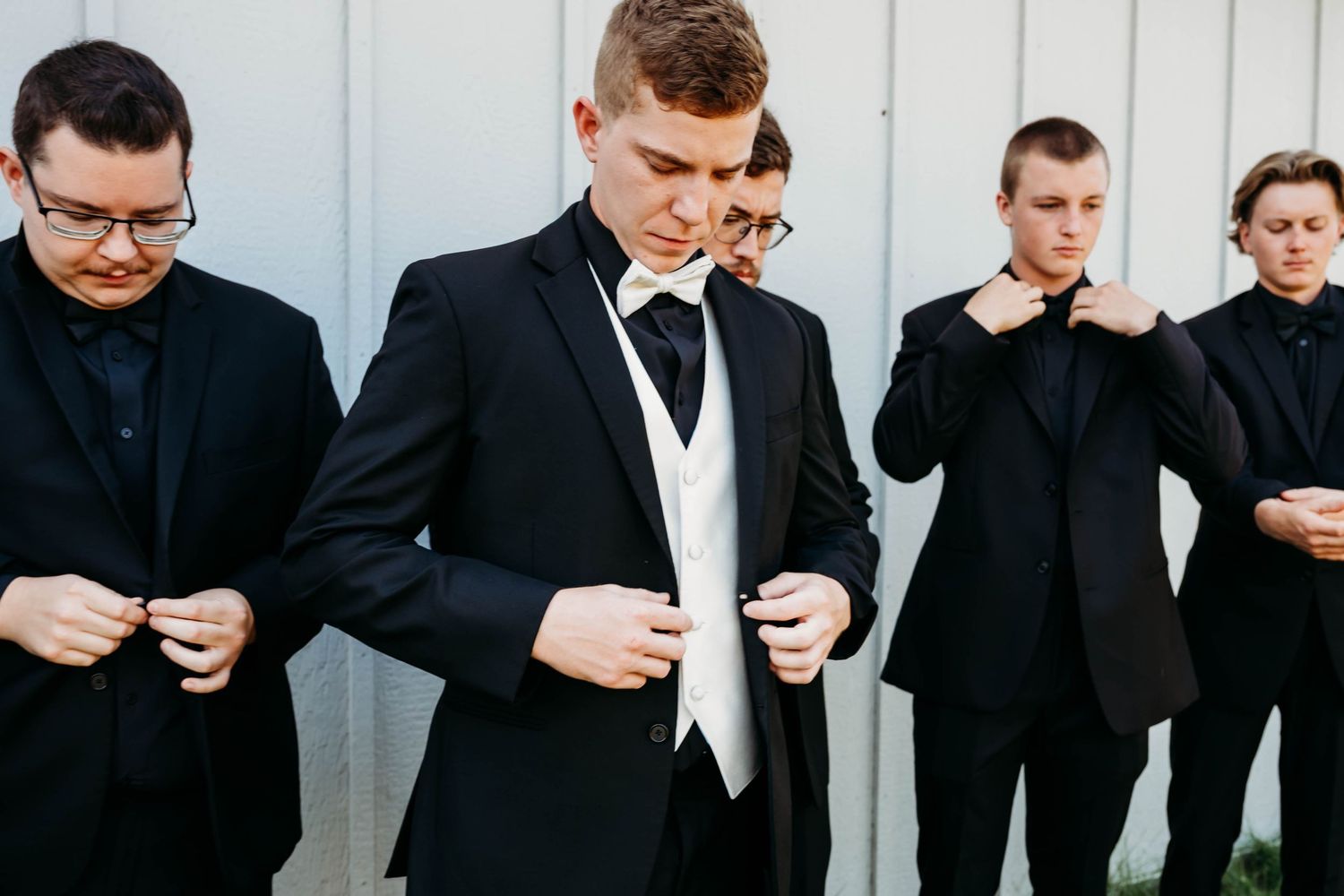 Groomsmen in black suits and tuxedos prepare for wedding ceremony against white wall background.