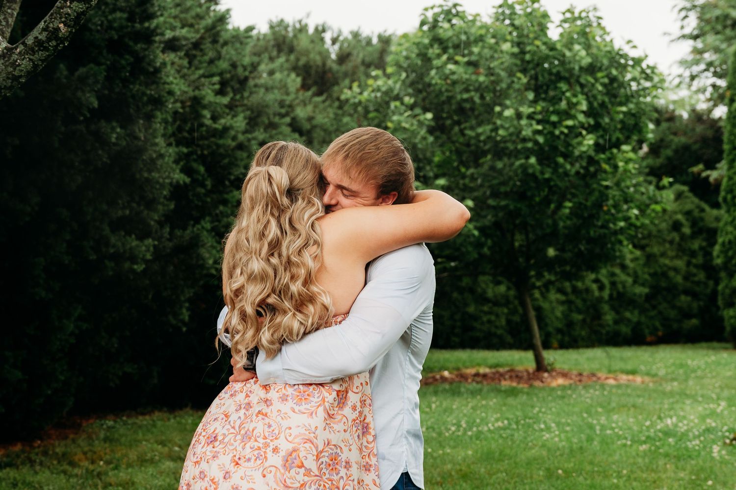 A couple embraces tenderly in a lush green garden, the woman wearing a floral dress with long curly hair.