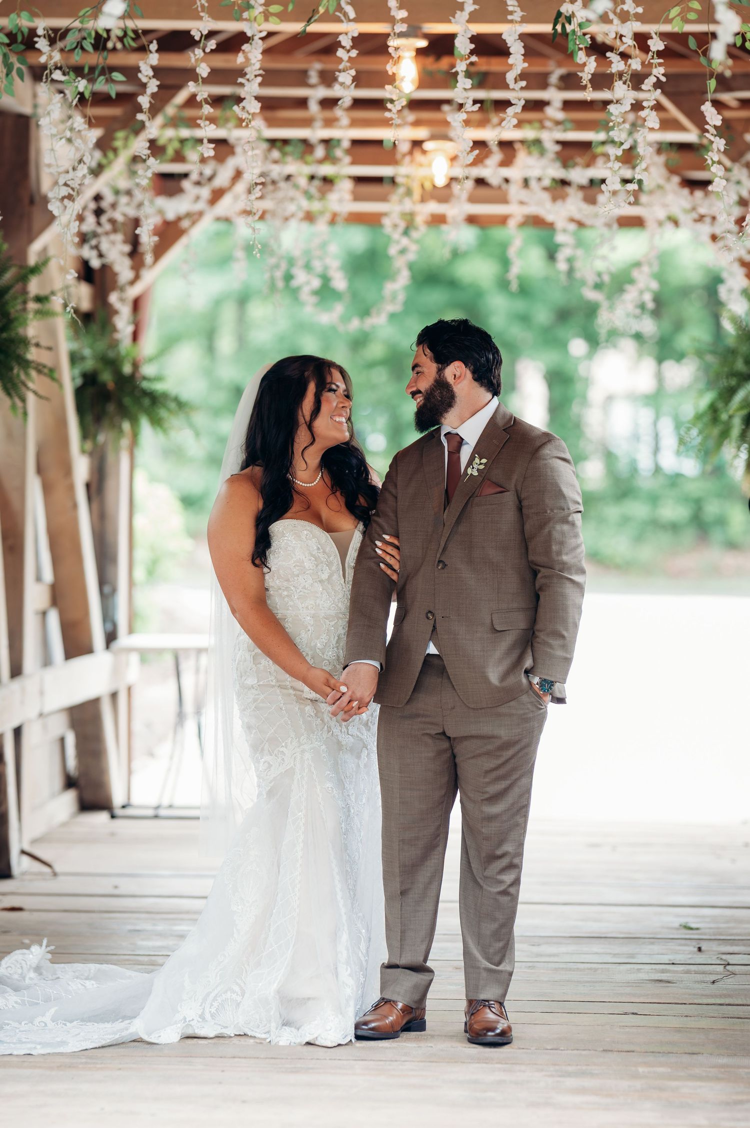 Smiling bride in lace gown and groom in tan suit share a joyful moment under a floral decorated pavilion.