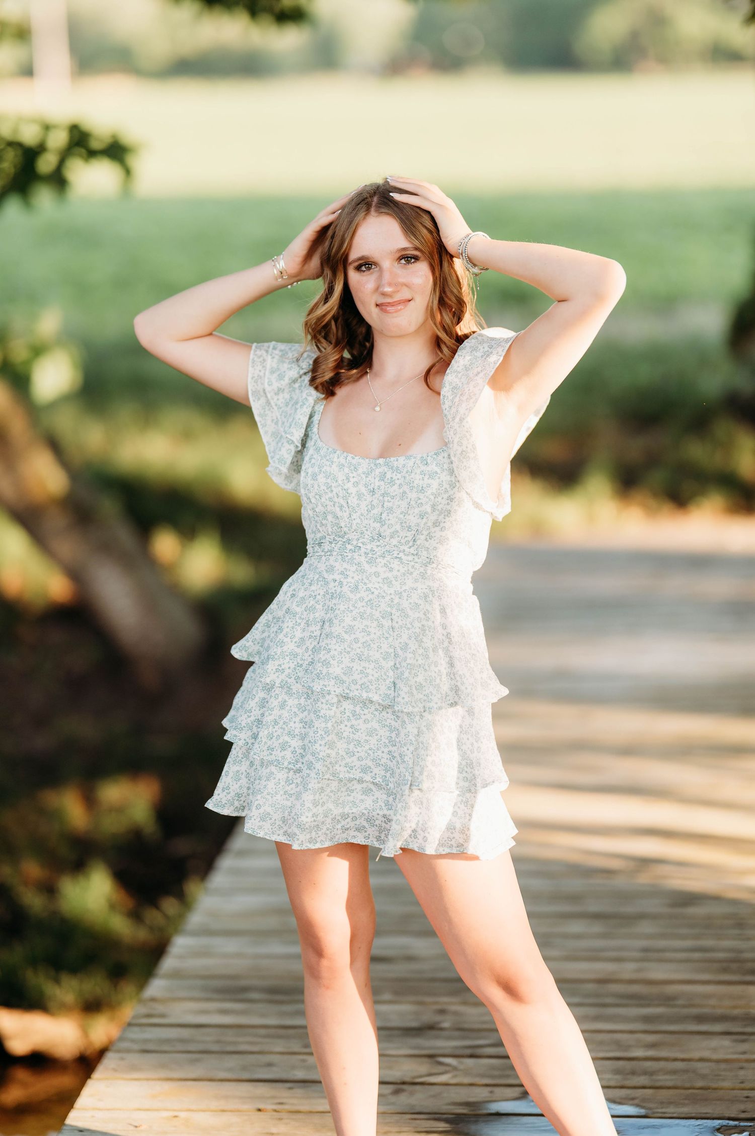 Young woman in floral white mini dress smiles on wooden boardwalk with hands in hair in warm outdoor lighting.
