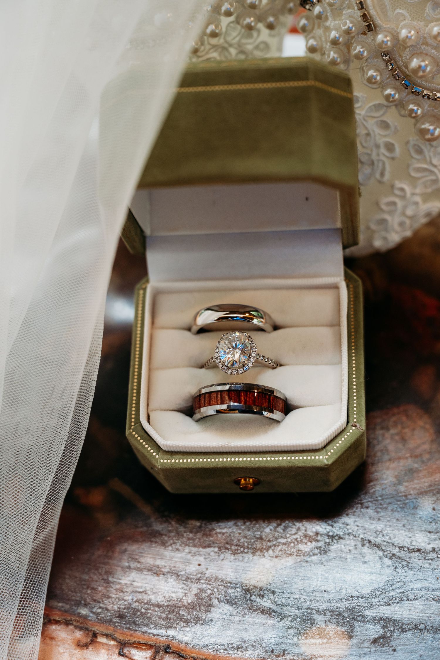 Three wedding rings displayed in green velvet box, featuring diamond halo engagement ring and wood inlay bands.