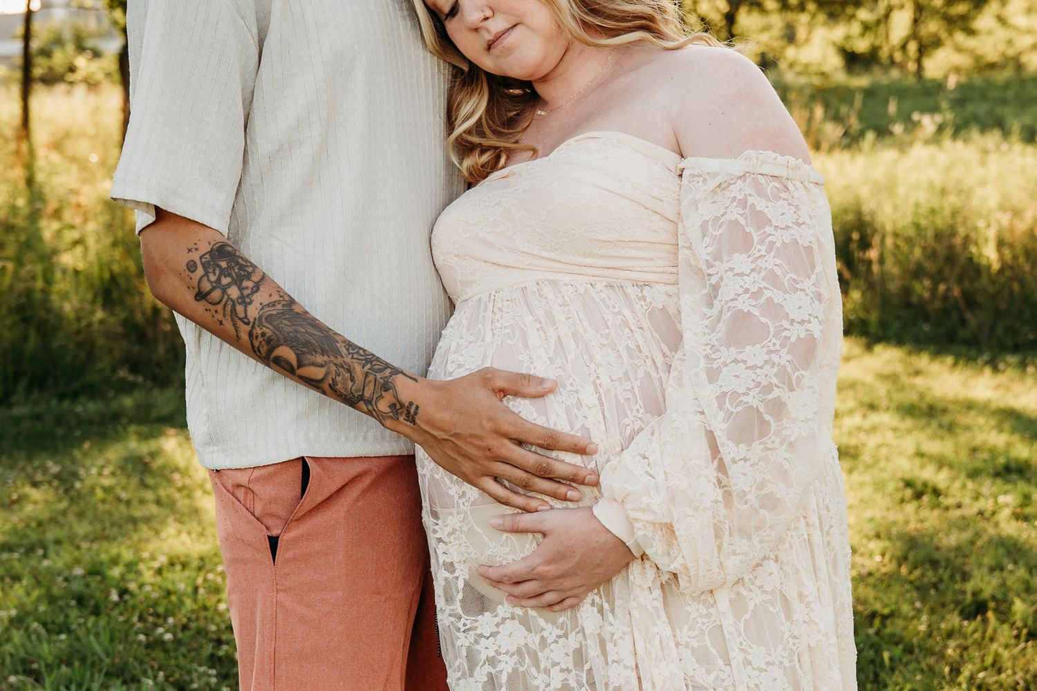 A pregnant person wearing a white lace dress stands with their partner in a sunlit outdoor setting.