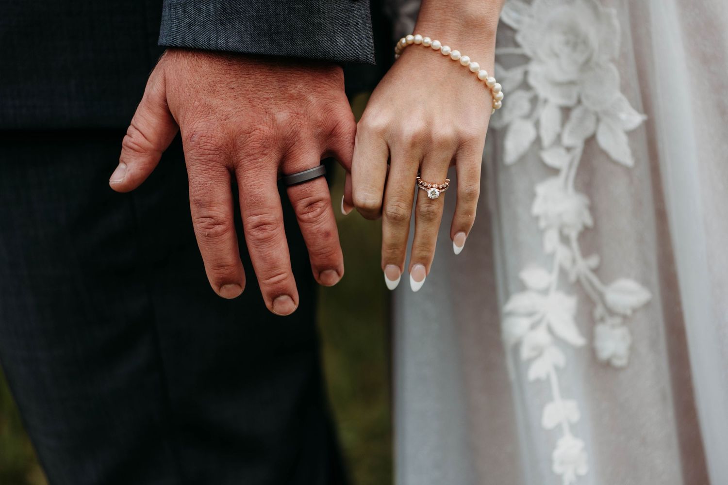 Close-up of two hands with wedding rings, one in a dark suit sleeve and one with pale manicured nails in white lace.