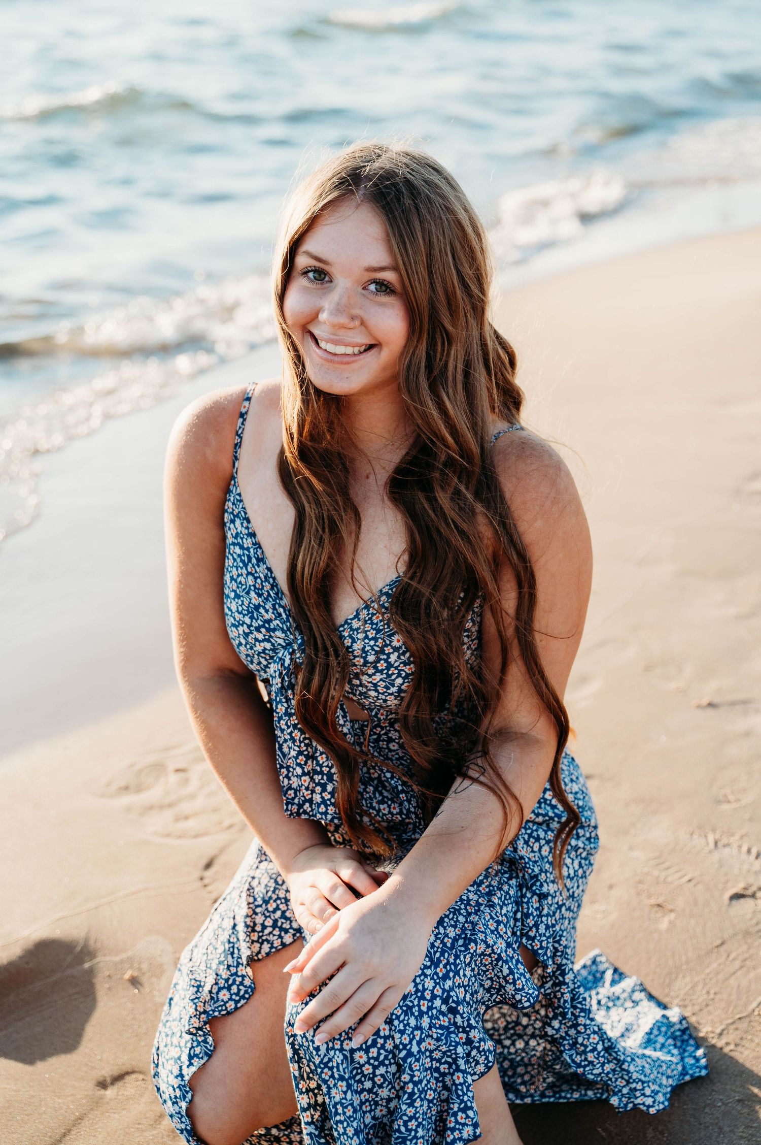 Woman in flowing blue floral dress sits smiling on sandy beach at sunset with ocean waves in background.