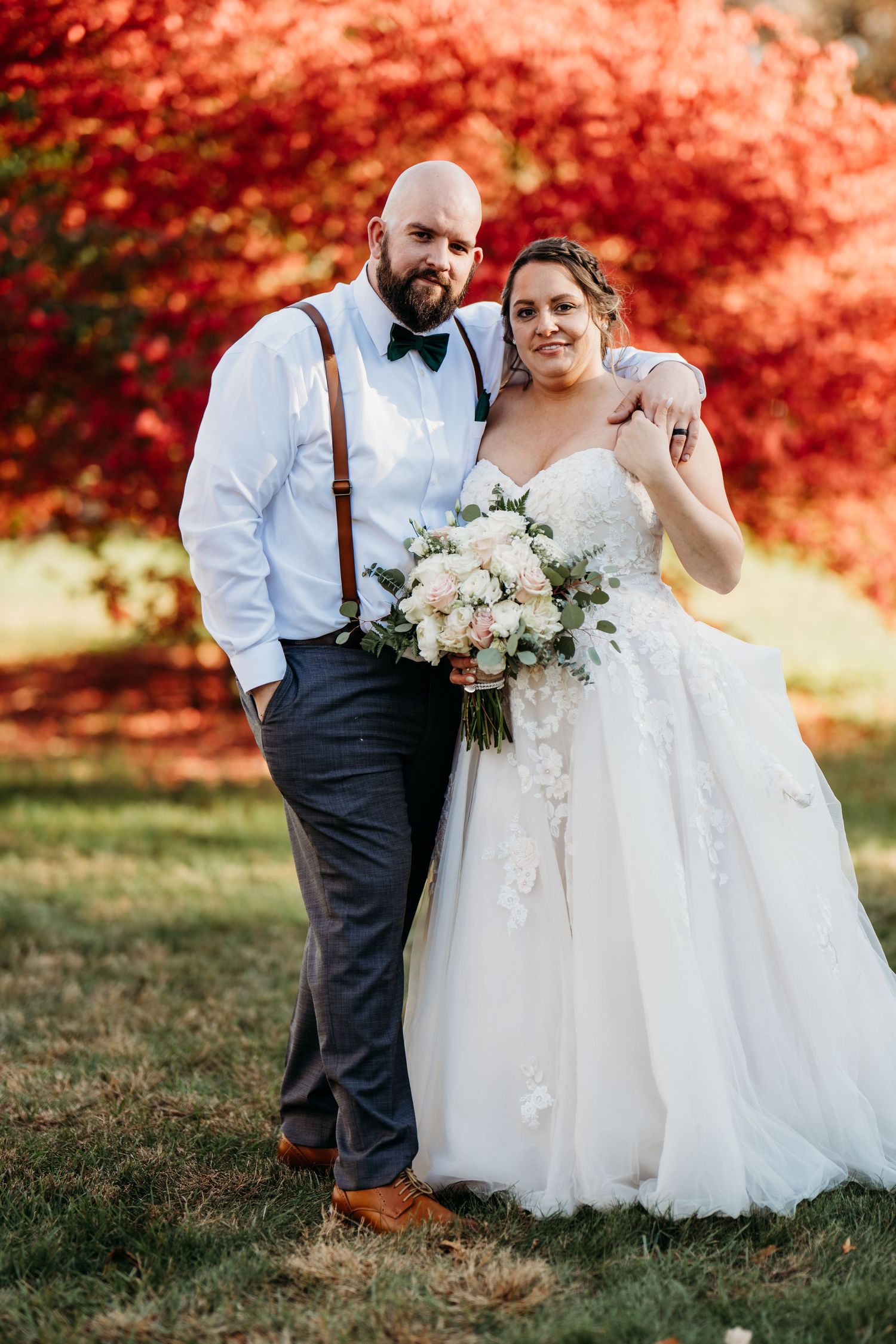 A couple poses during their fall wedding ceremony against vibrant red foliage while holding a white and blush bouquet.