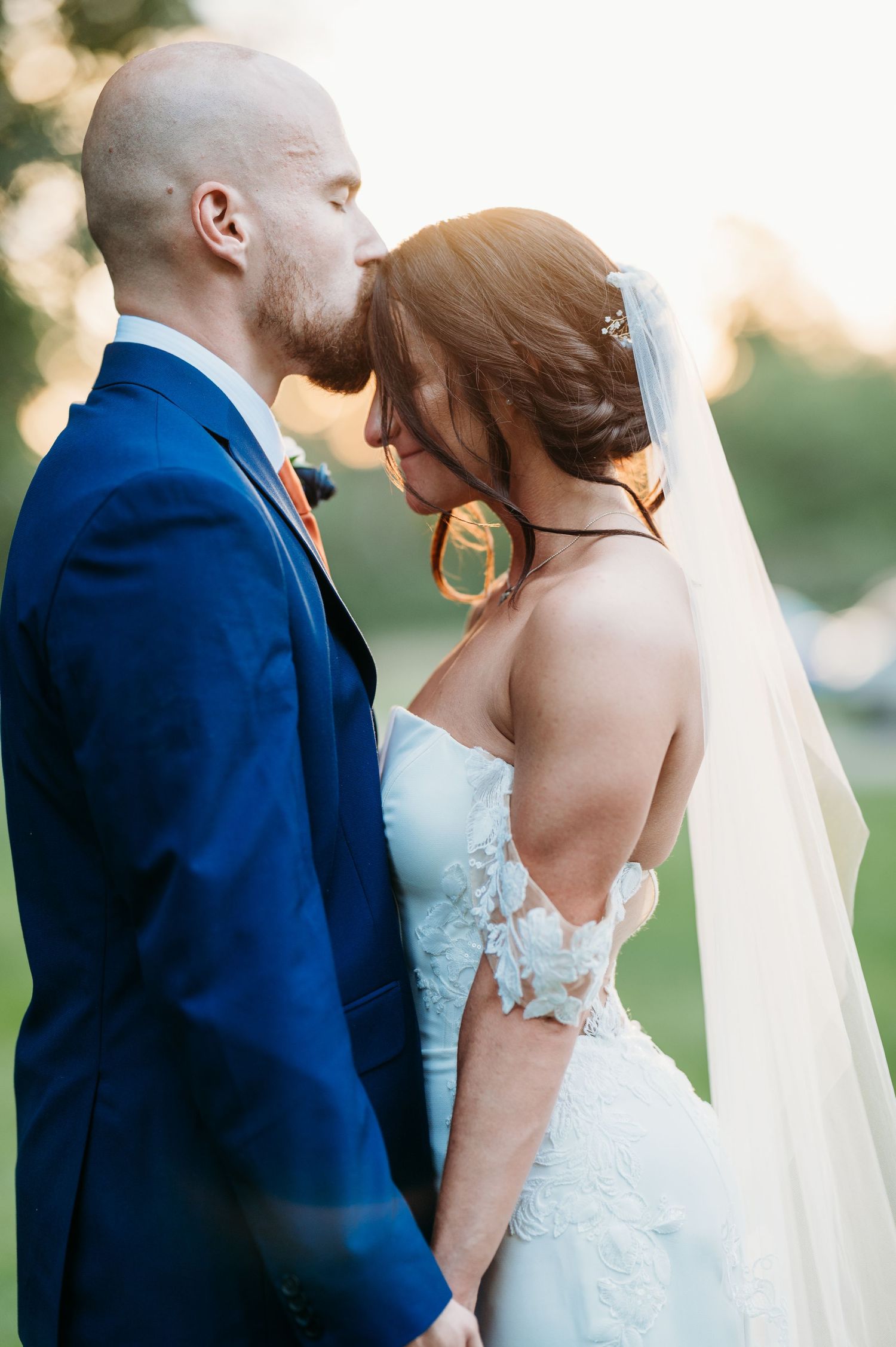 Romantic wedding couple in navy suit and white lace dress share intimate moments during sunset outdoor photo session.