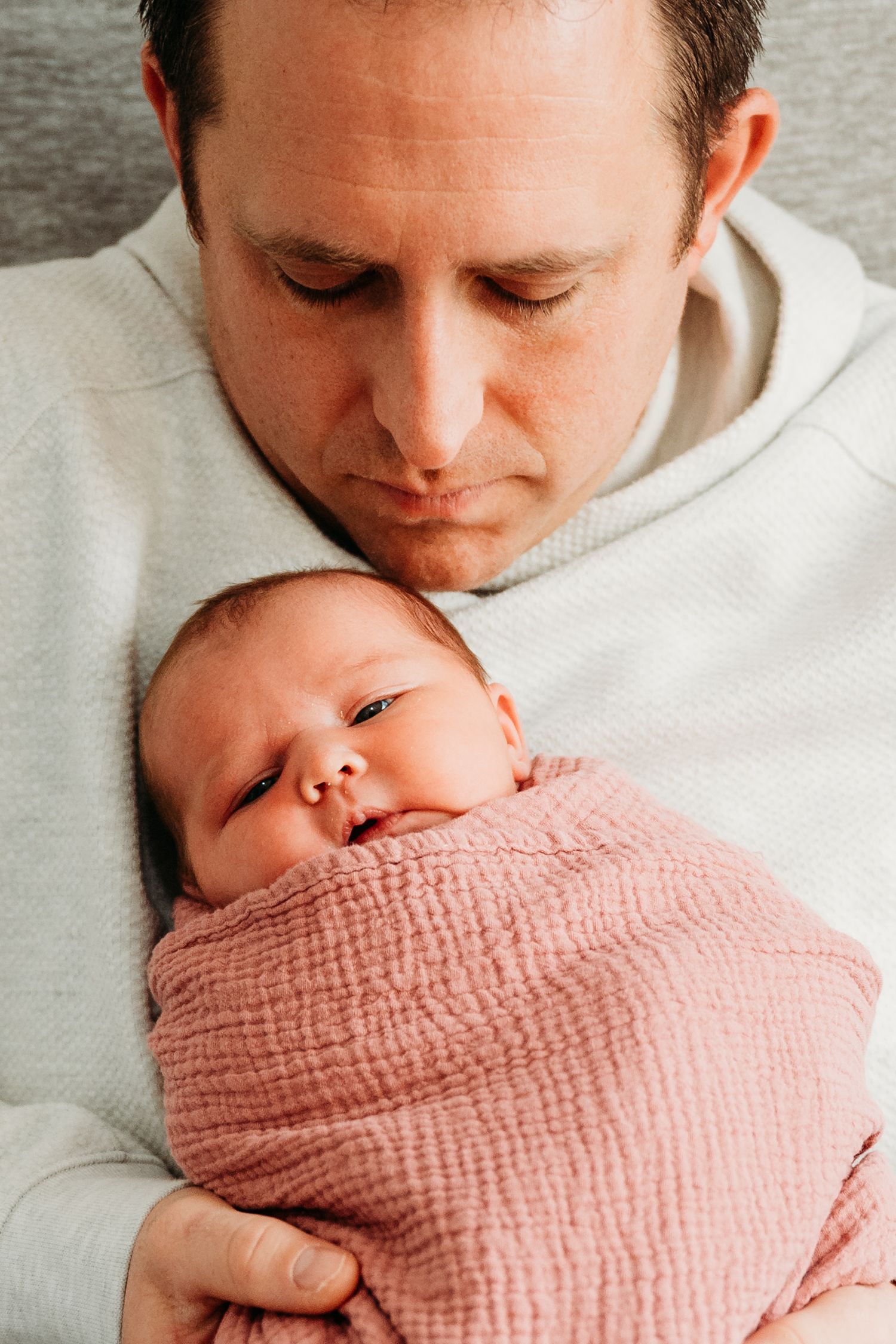 A parent holds a sleeping newborn baby wrapped in a soft pink muslin swaddle blanket against a light gray background.