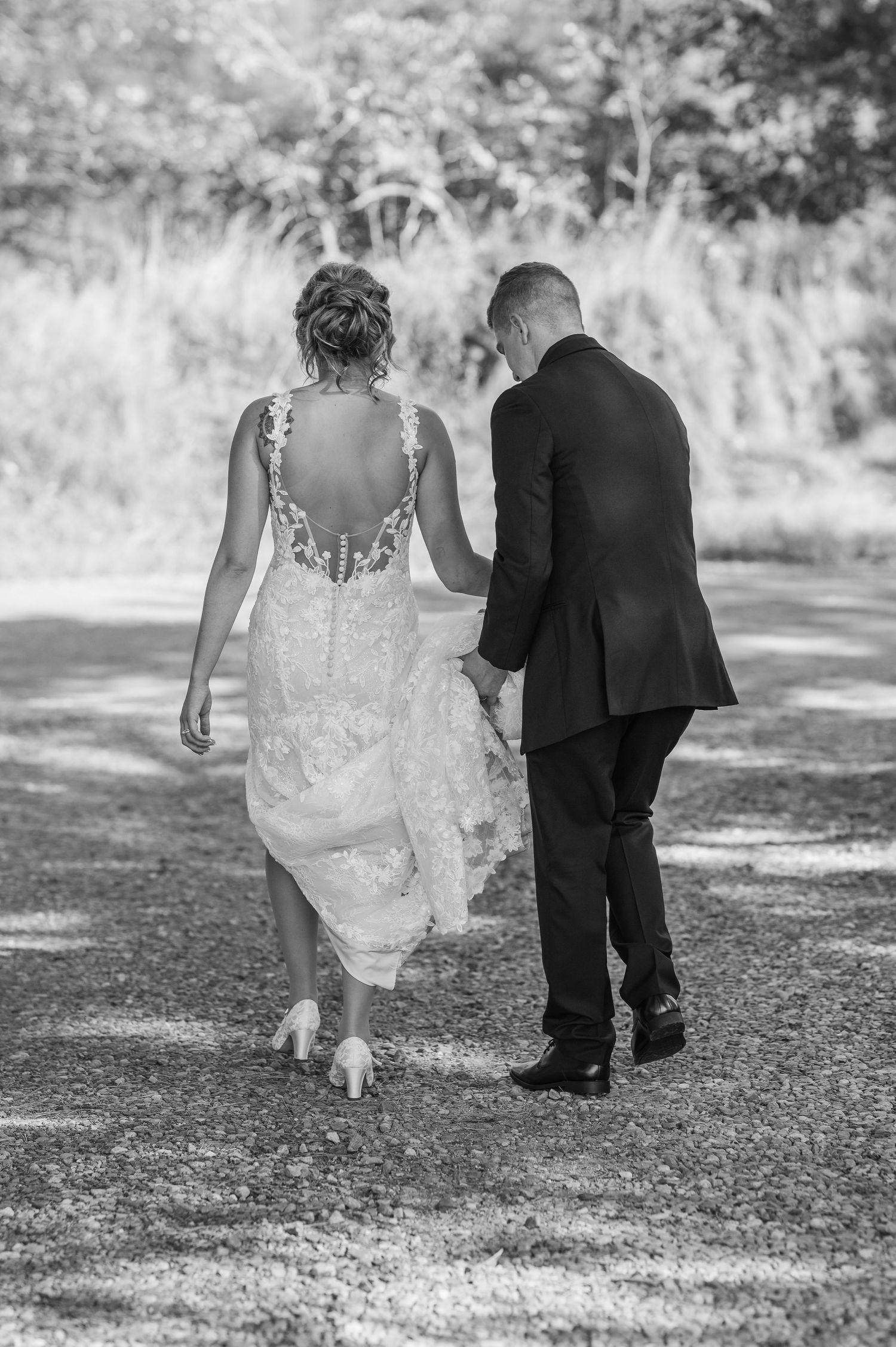 Black and white photo of couple walking together on gravel path in wedding attire.