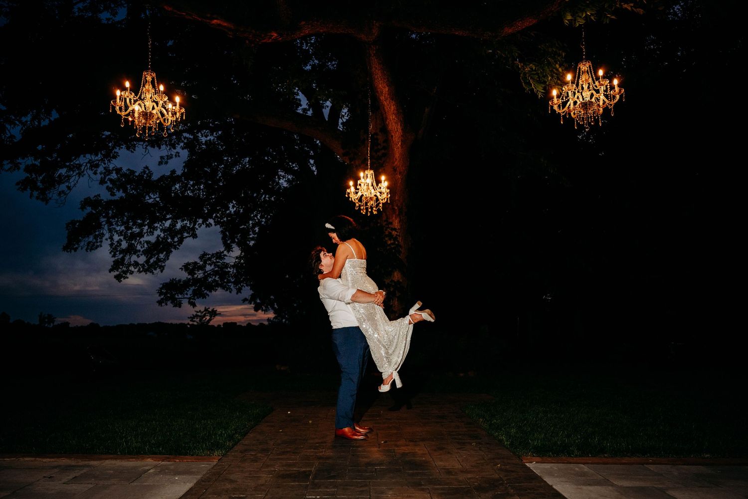 Couple dancing under chandeliers hanging from a tree at dusk during an outdoor wedding celebration.