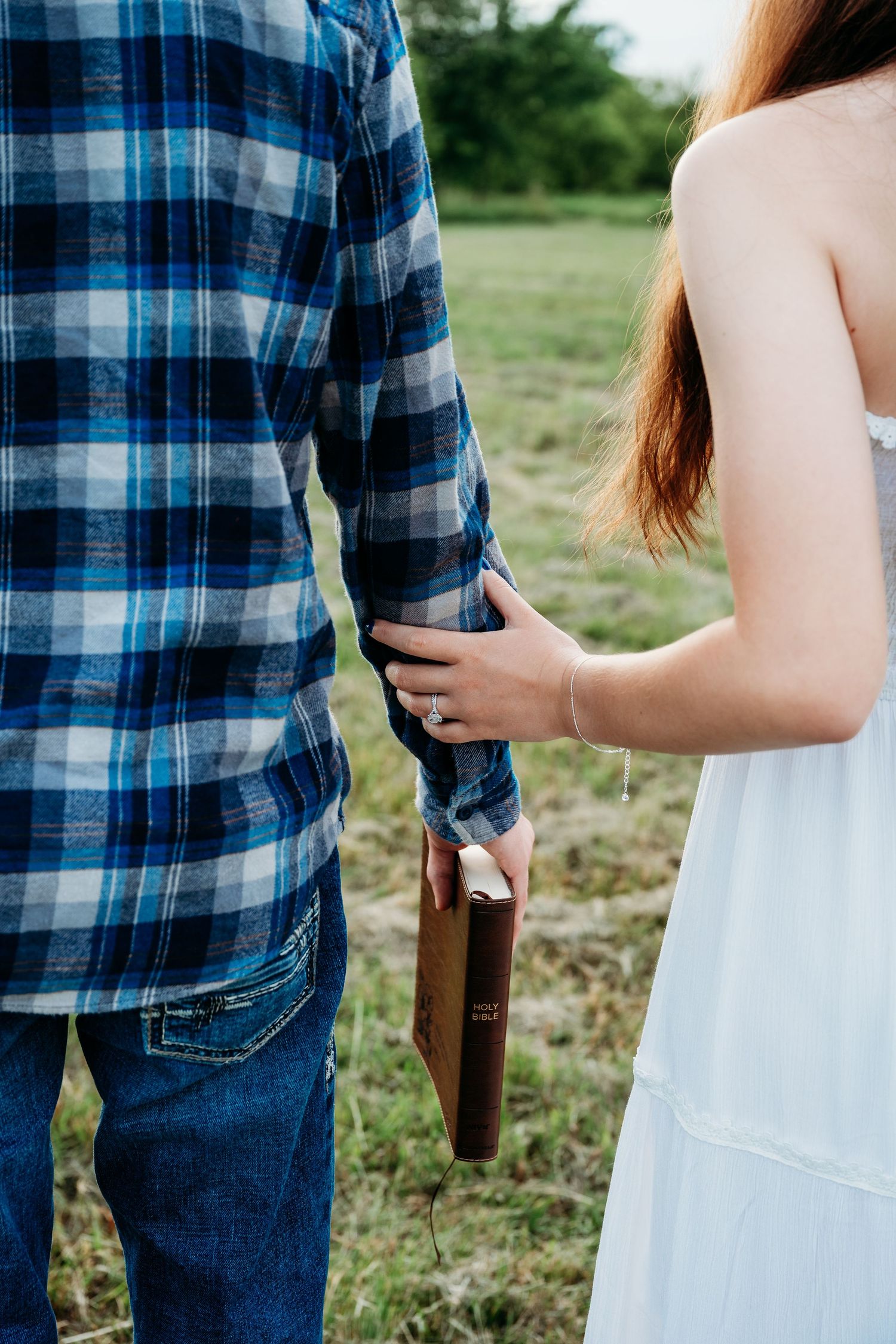 Couple holding hands and a Bible together in a field, woman wearing a ring in a white dress.
