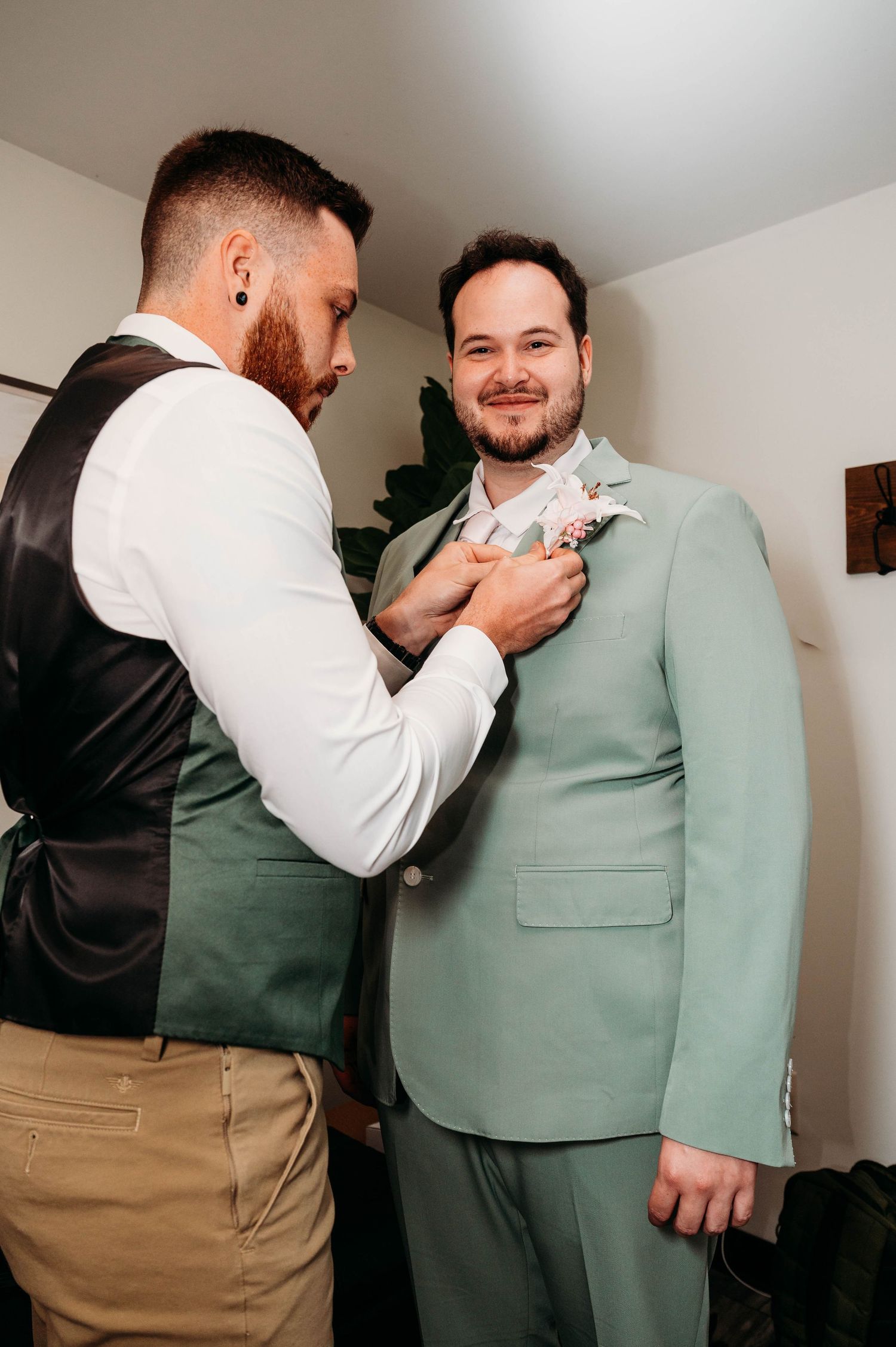 Groom in mint green suit gets boutonniere pinned by groomsman in vest before wedding ceremony.