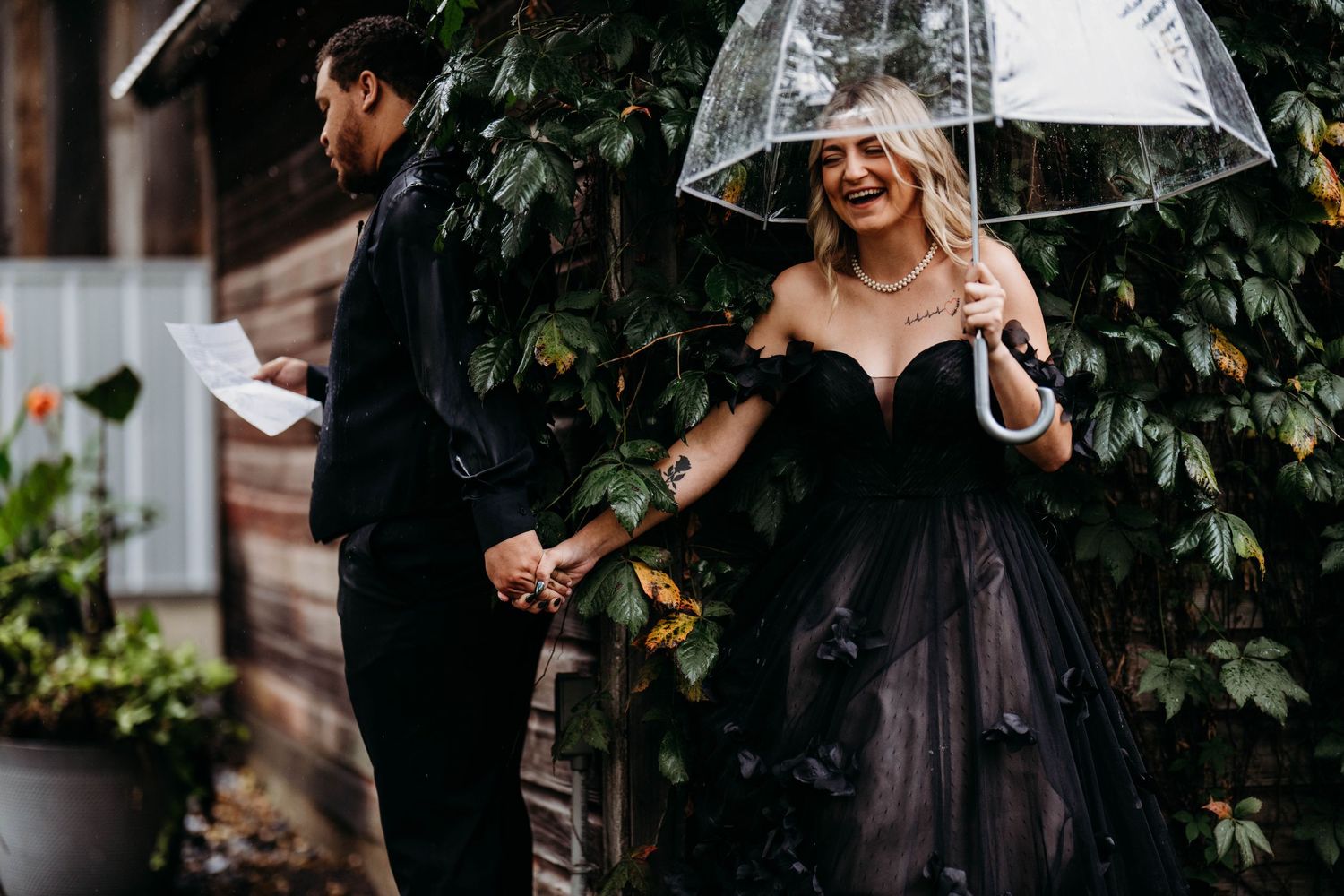 A couple in elegant black attire holding hands while one holds a clear umbrella near a garden wall.