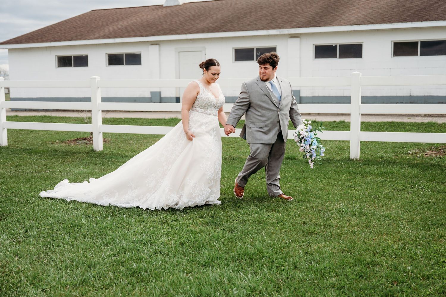 Couple in wedding attire walking together on green grass lawn in front of white farm fence and barn.