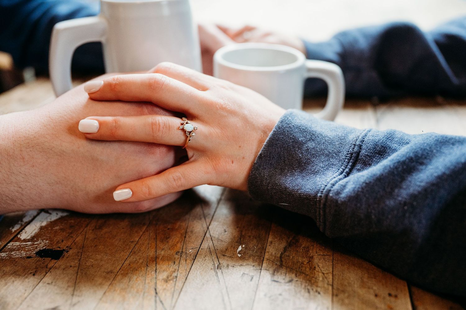 Hands with engagement ring rest on wooden table next to white coffee mugs.