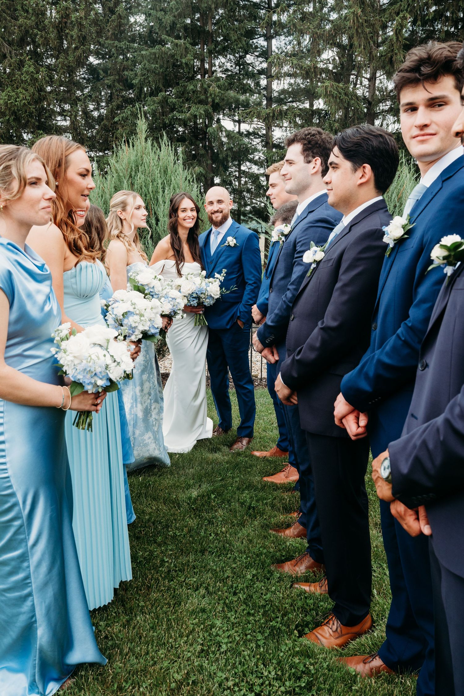 Wedding party lines up outdoors wearing coordinated blue dresses and navy suits with floral bouquets.