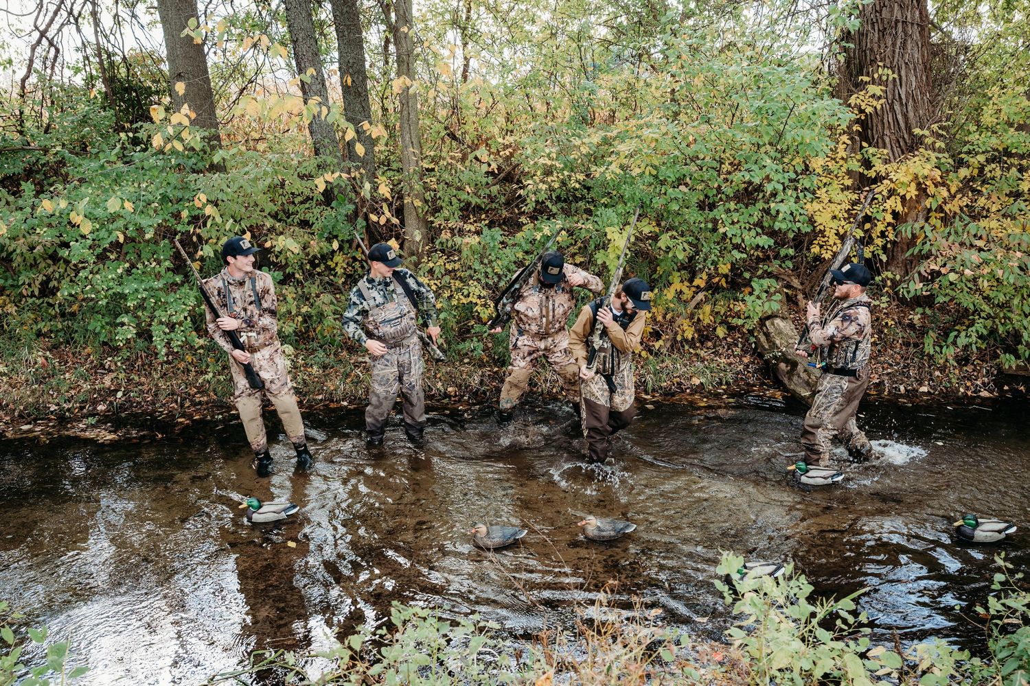 Group of hunters in camouflage gear crossing a shallow creek during autumn season.