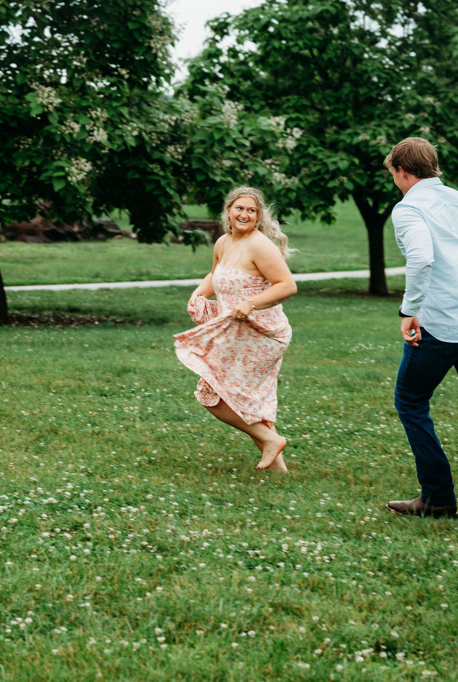 Joyful woman in floral dress twirling barefoot on green grass while her partner reaches for her hand.