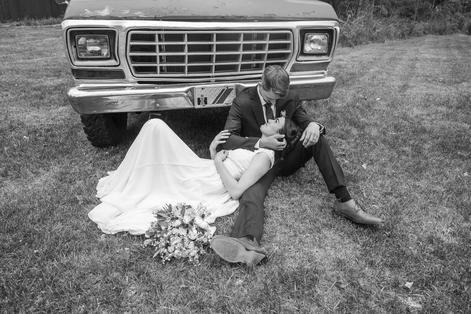 Black and white photo of a bride and groom sitting together against a vintage truck, sharing a romantic moment.