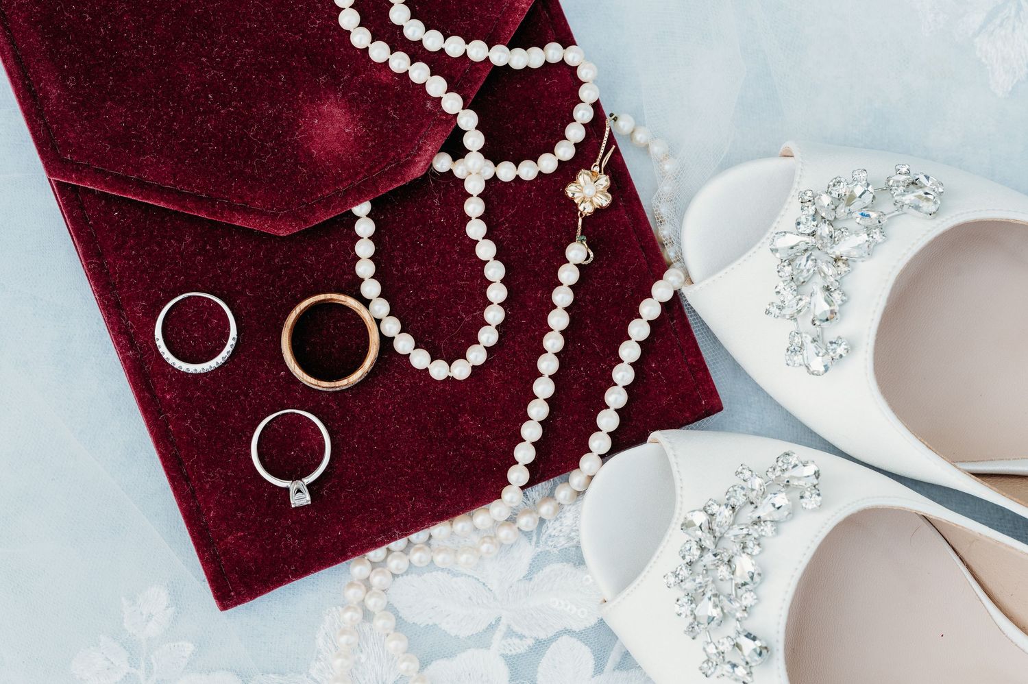 Elegant wedding flat lay with pearl necklace, gold and silver rings on red velvet, and white jeweled bridal shoes.