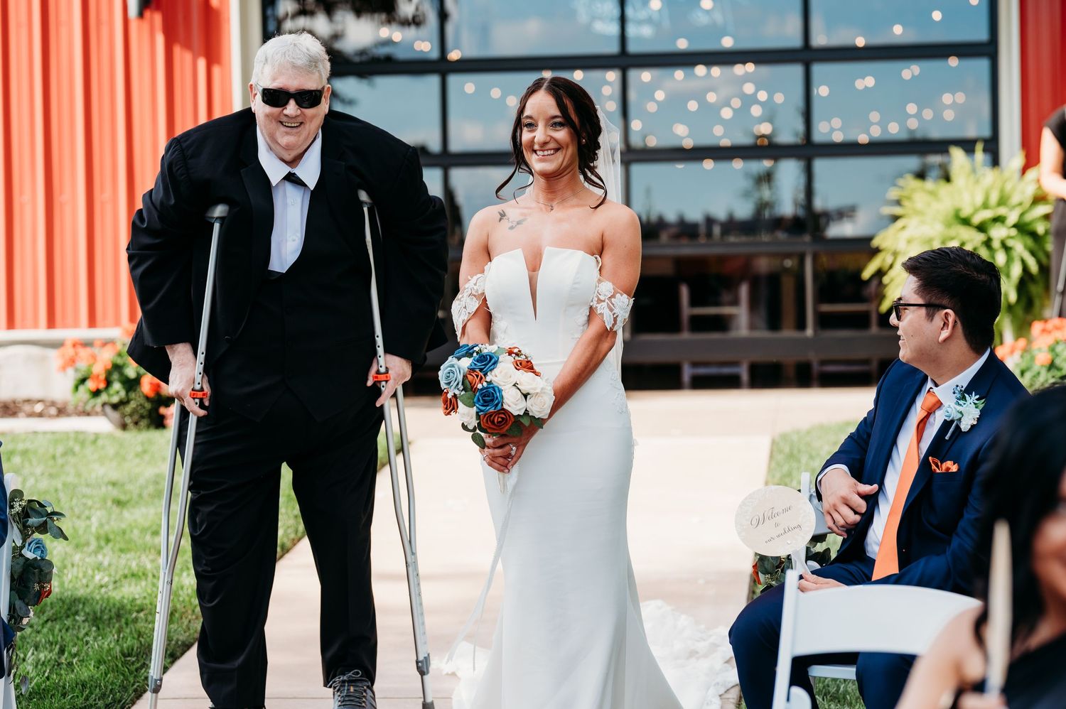 Bride in white gown smiles while walking with man on crutches at outdoor wedding ceremony.