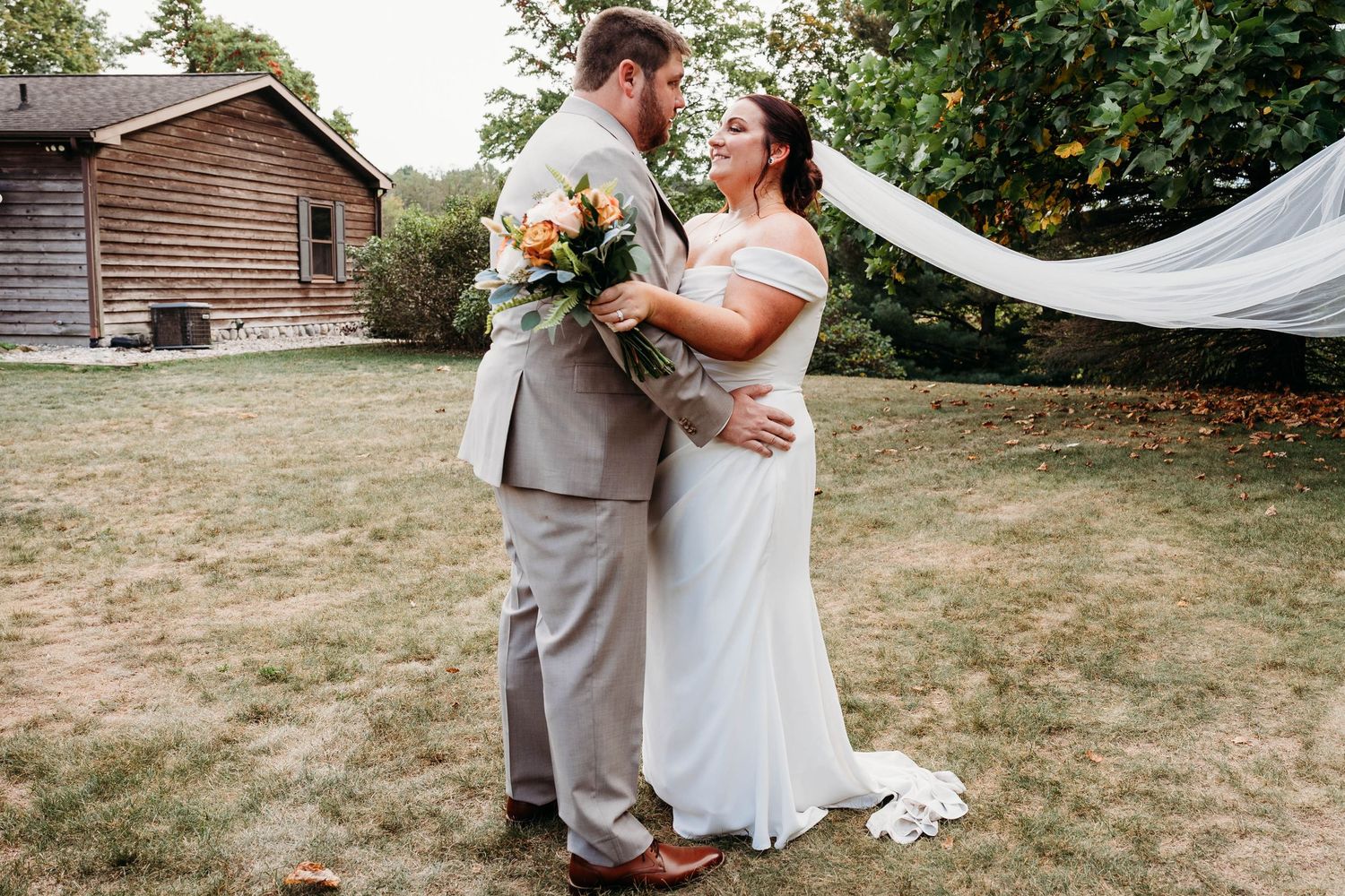 Bride and groom share a romantic moment outdoors, her veil flowing in the wind, holding a colorful bouquet near a rustic barn.