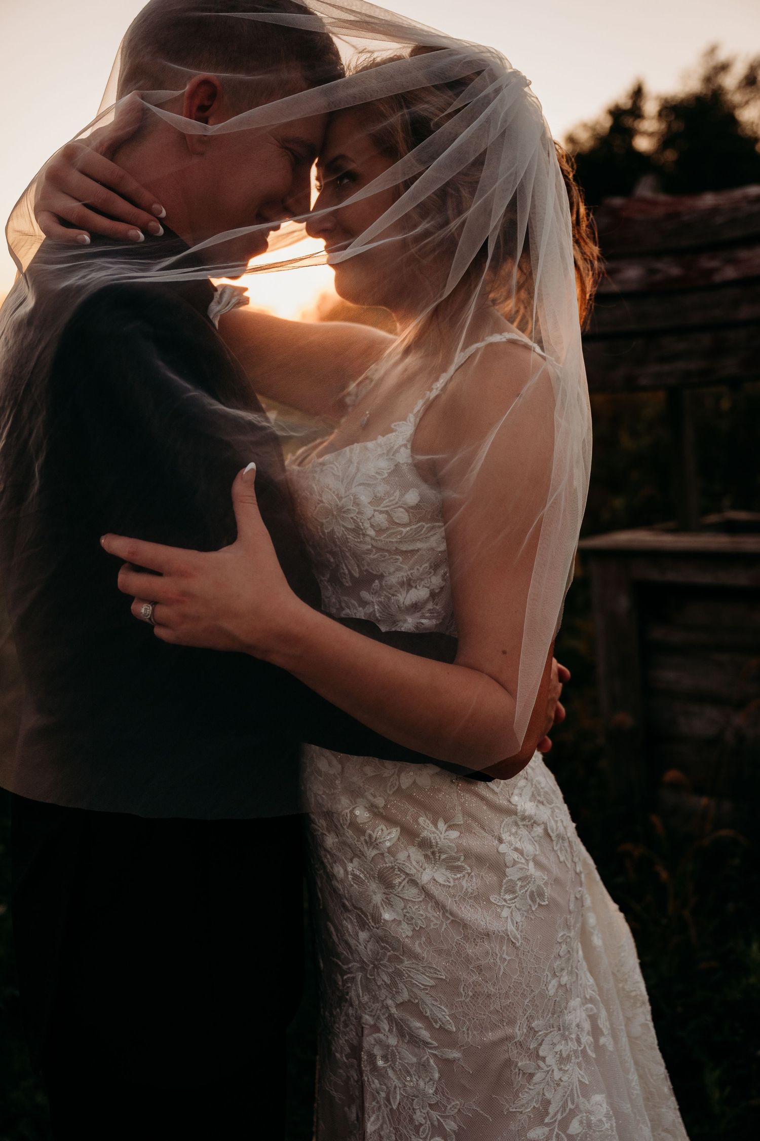 Bride and groom embracing under wedding veil at golden sunset, romantic backlit portrait.