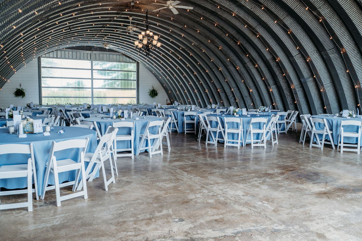 Elegant wedding reception in a rustic quonset barn with blue tablecloths and white folding chairs under a chandelier.