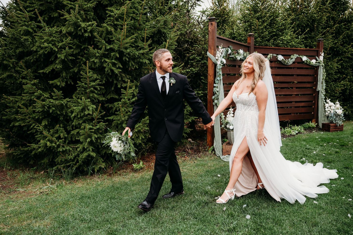Bride and groom walk hand-in-hand on green lawn with evergreen trees and floral arch backdrop.
