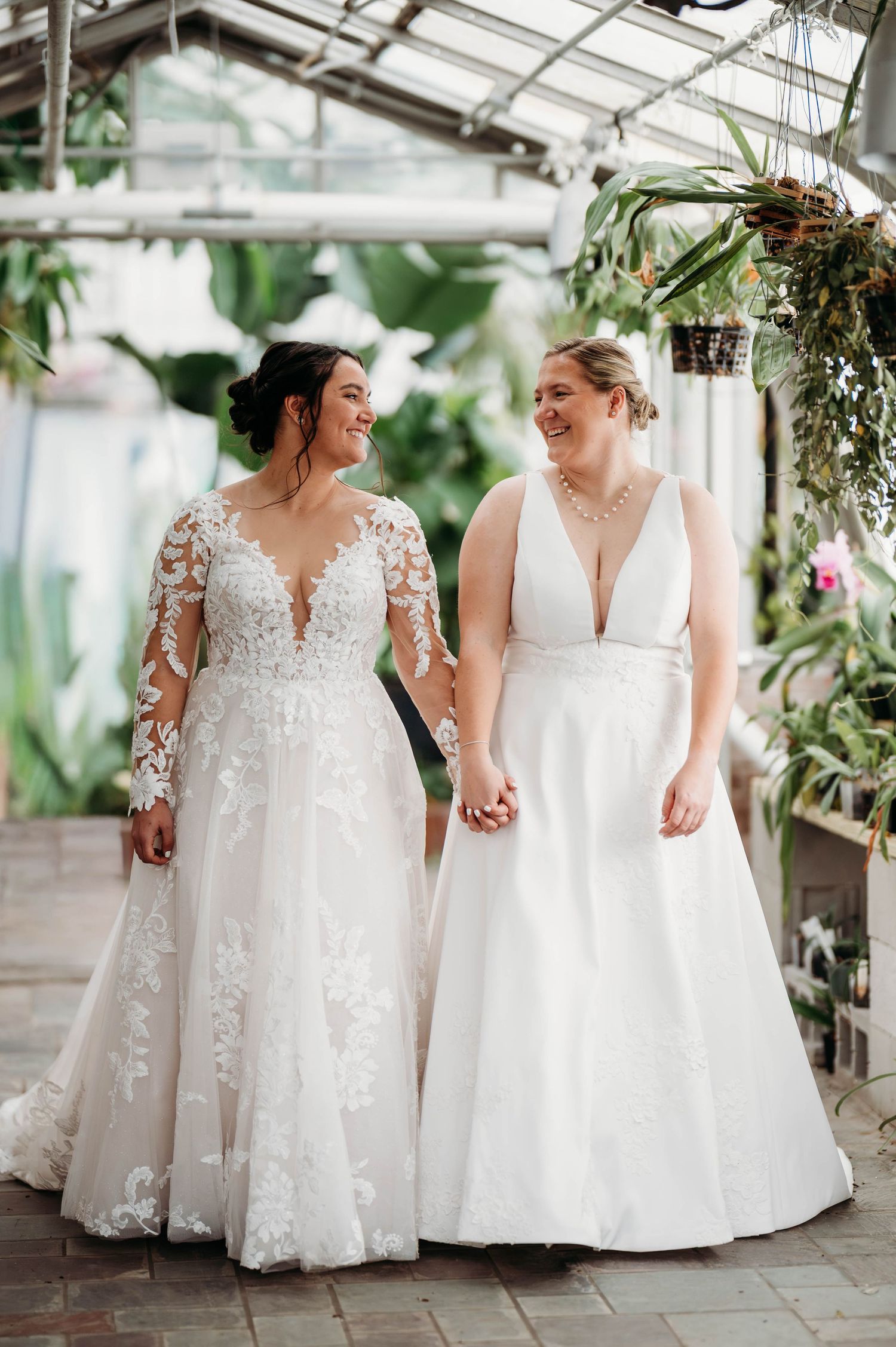 Two people in elegant white wedding gowns hold hands while walking together in a greenhouse filled with greenery and natural light.
