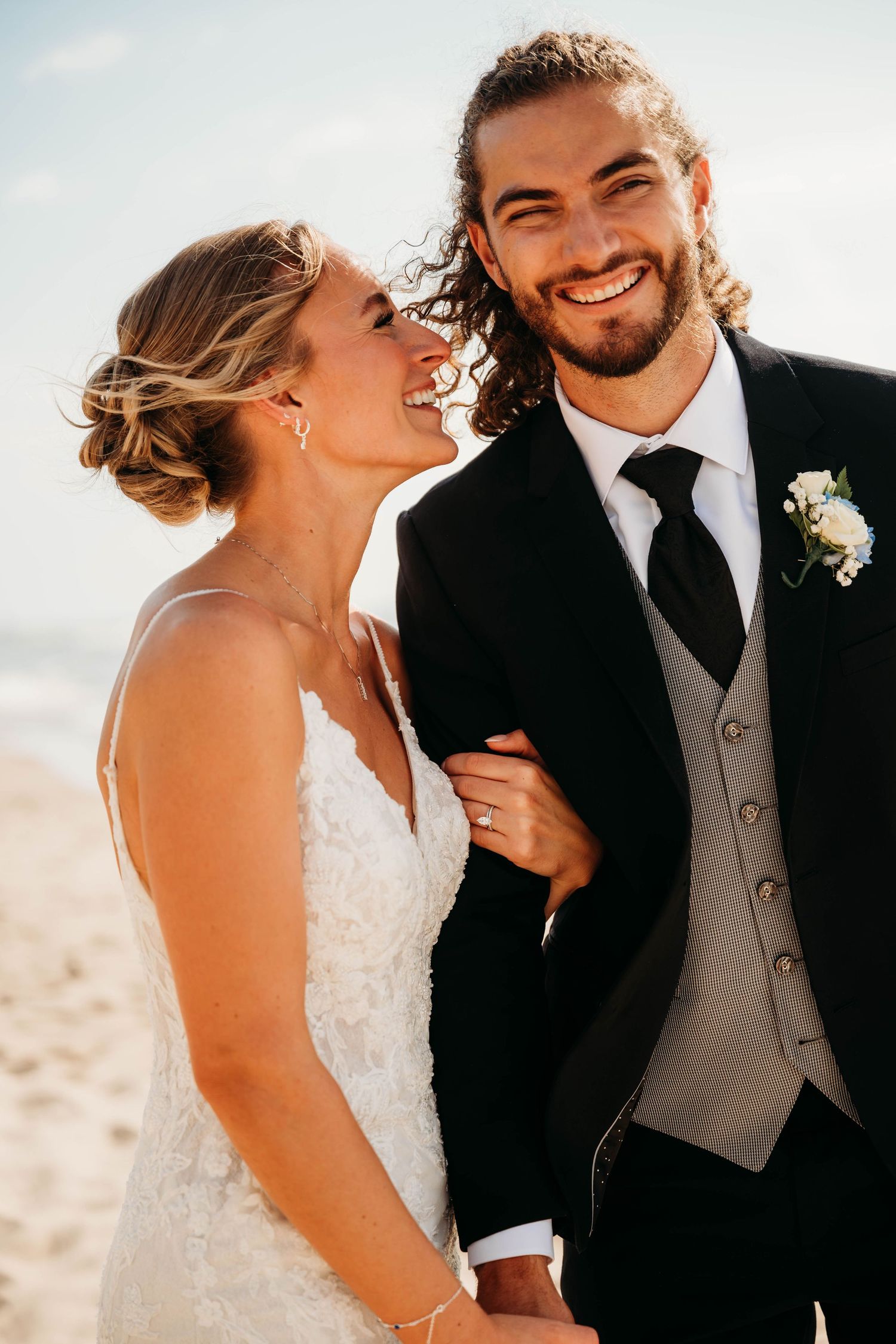 A smiling bride in white lace gown and groom in black tuxedo pose together on a sunny beach.