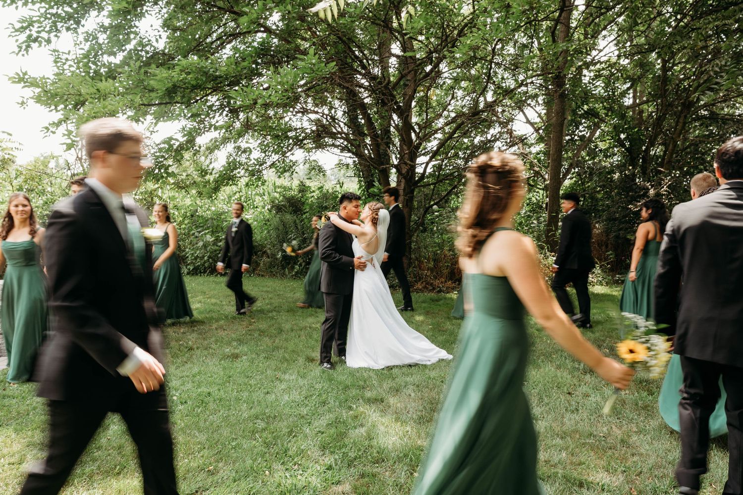 Bride and groom share a kiss surrounded by wedding party in green dresses and black tuxedos on a lush garden lawn.