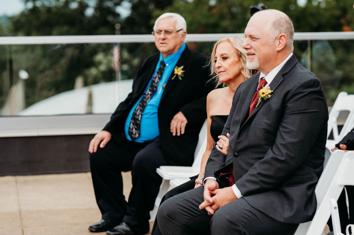 People in formal attire gather outdoors for a wedding ceremony with chairs arranged in rows on a sunny day.