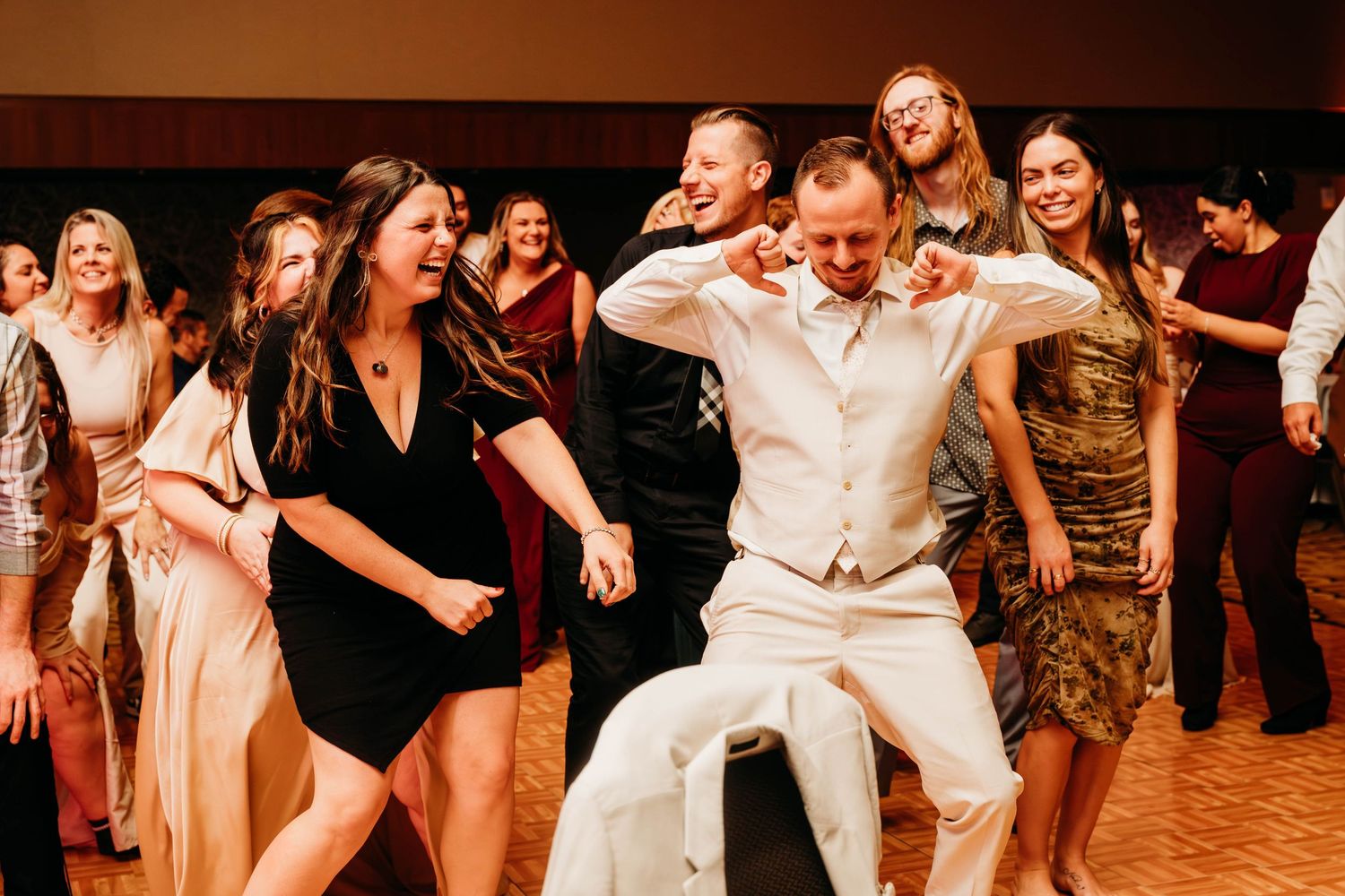 Wedding party guests dancing and celebrating on a dance floor at a reception in warm lighting.