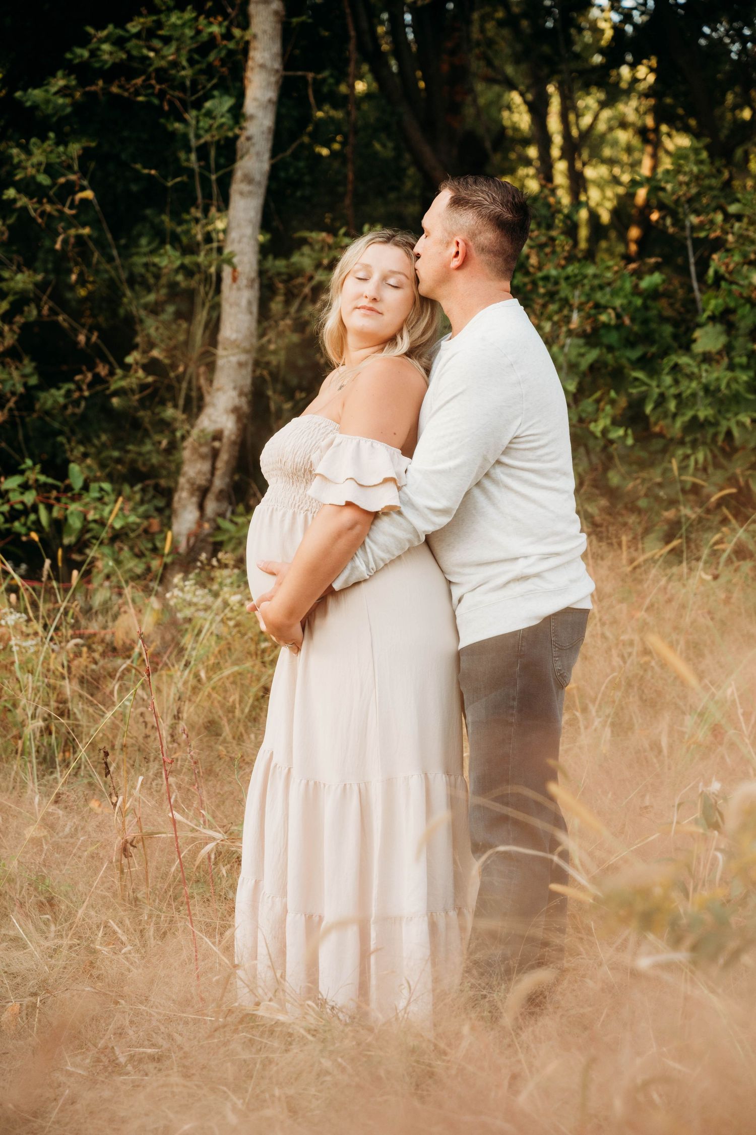 A romantic couple in light colored clothing embrace in a sunlit meadow with tall grass and trees in the background.