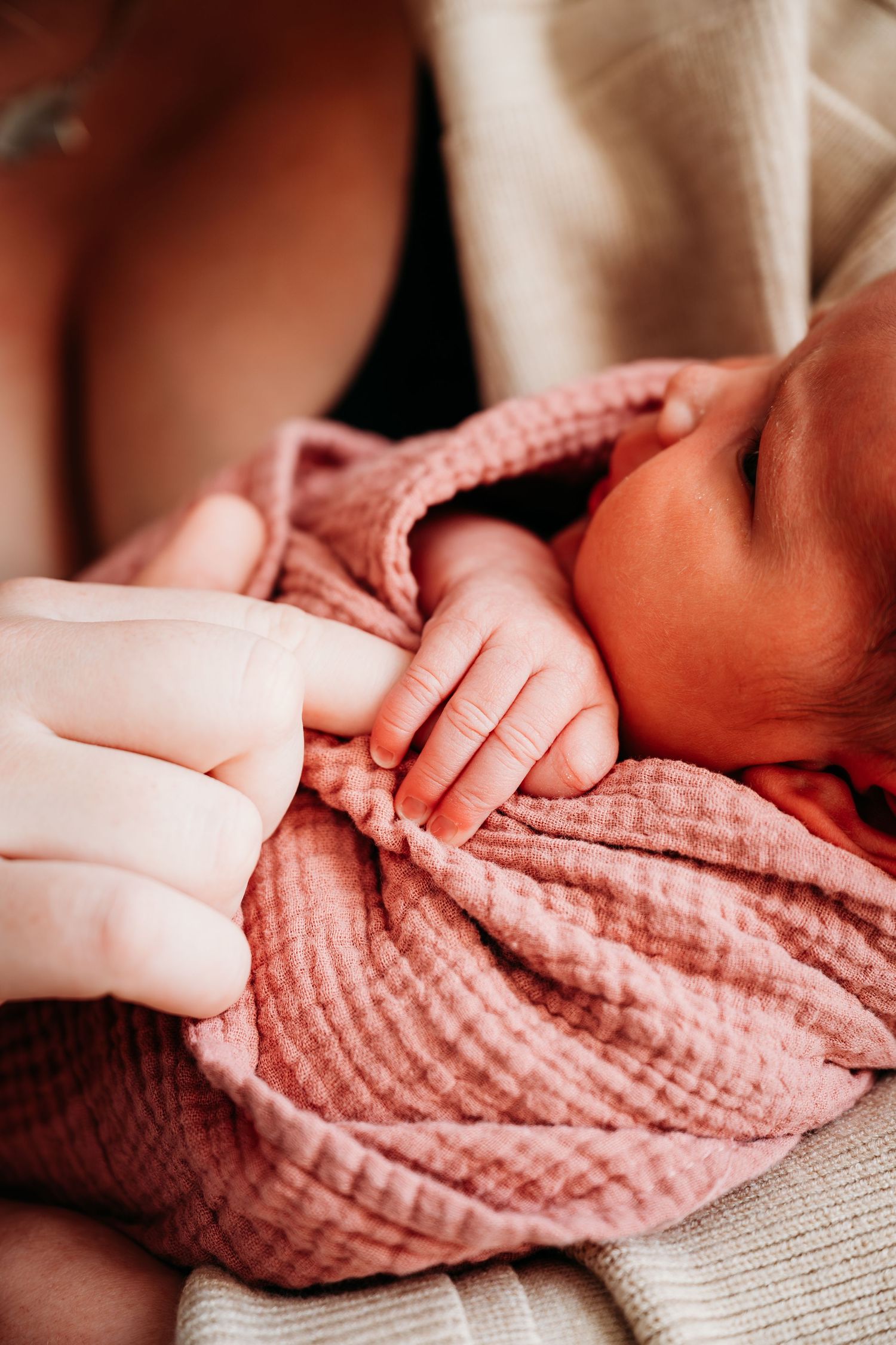 A tiny baby hand grasps an adult finger while wearing a soft pink muslin swaddle blanket.