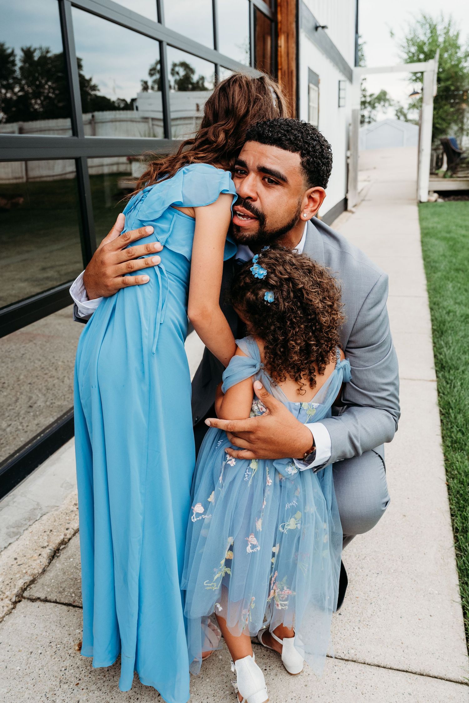 A parent in a gray suit lovingly embraces a child in a light blue dress during an emotional outdoor moment.