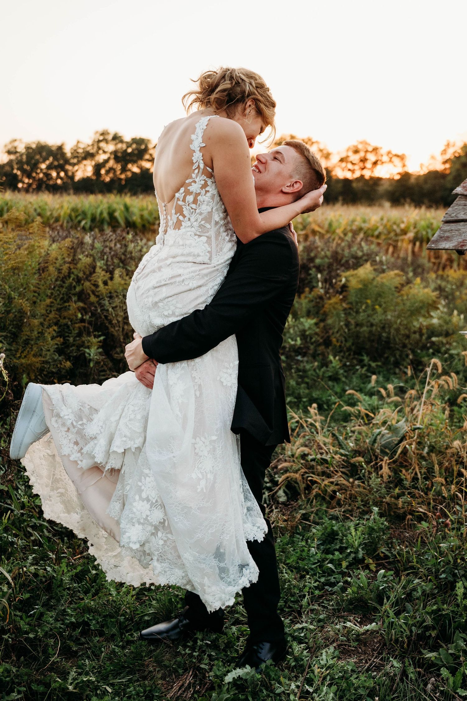 A romantic sunset moment in a wildflower field as a couple embraces, with the bride's lace dress flowing in the breeze.