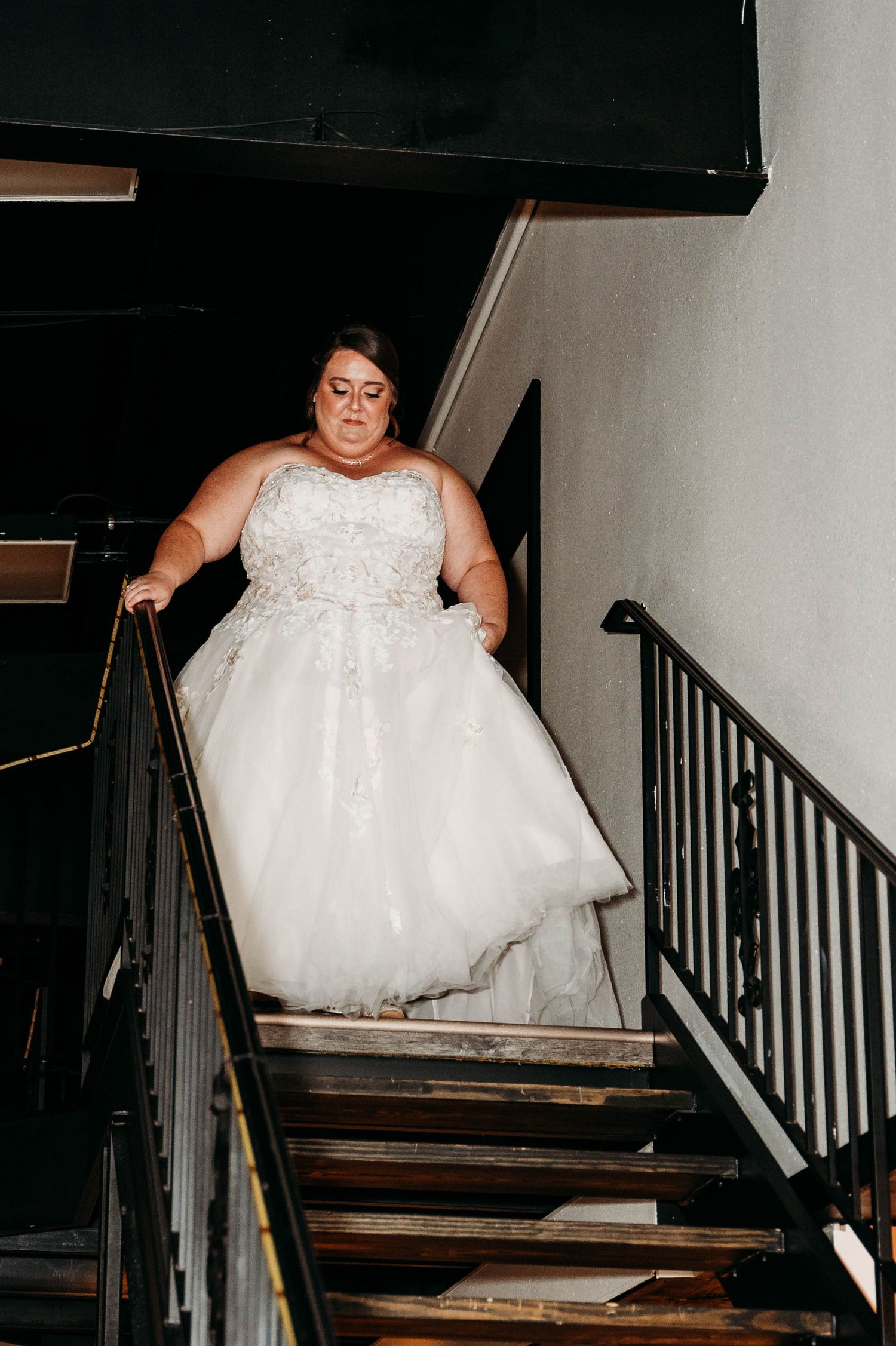 A person in a white lace wedding dress descends a wooden staircase with black railings.