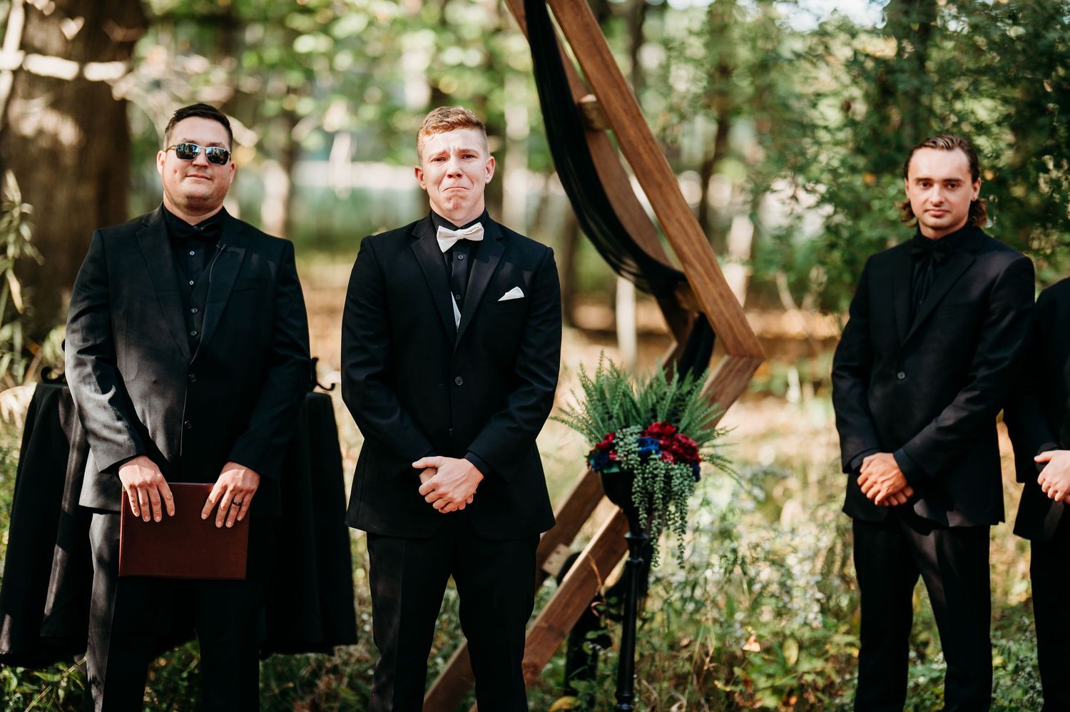 Three groomsmen in matching black suits and sunglasses pose together outdoors near rustic wooden decor.
