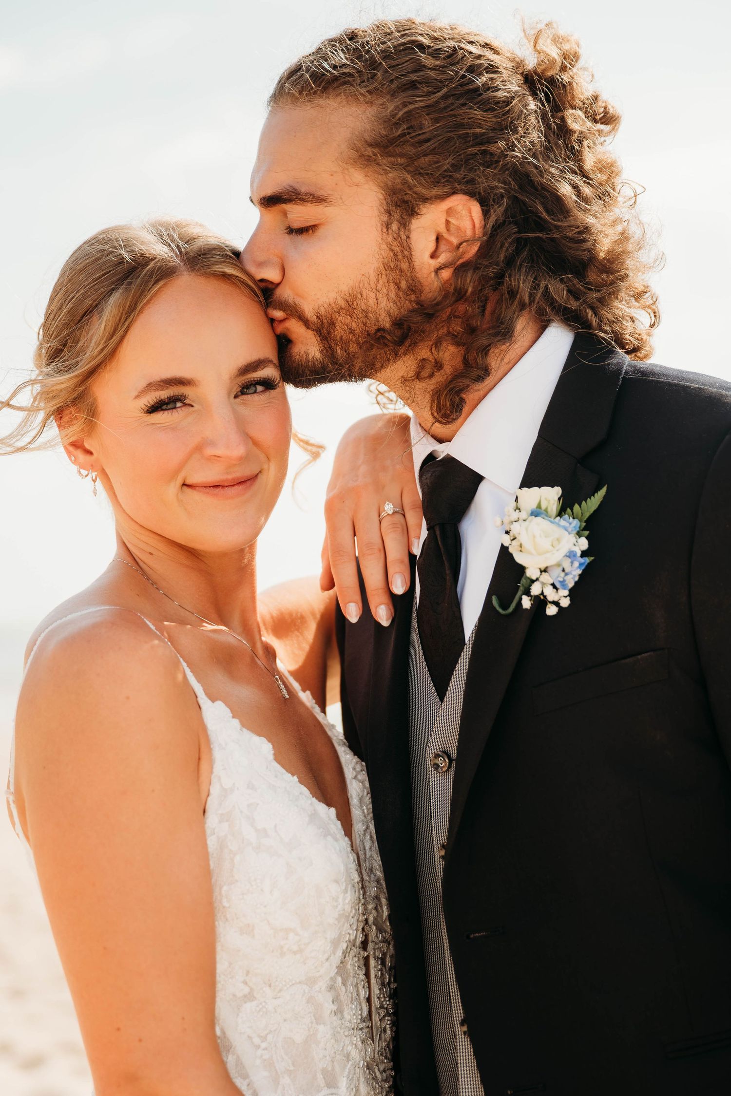 Groom kisses brides temple as she smiles warmly, both in wedding attire on a sunlit beach.