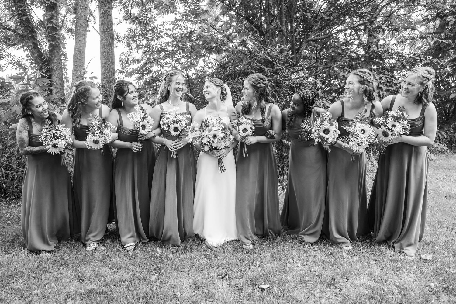 Black and white photo of a bride surrounded by bridesmaids in long dresses holding bouquets, laughing outdoors.