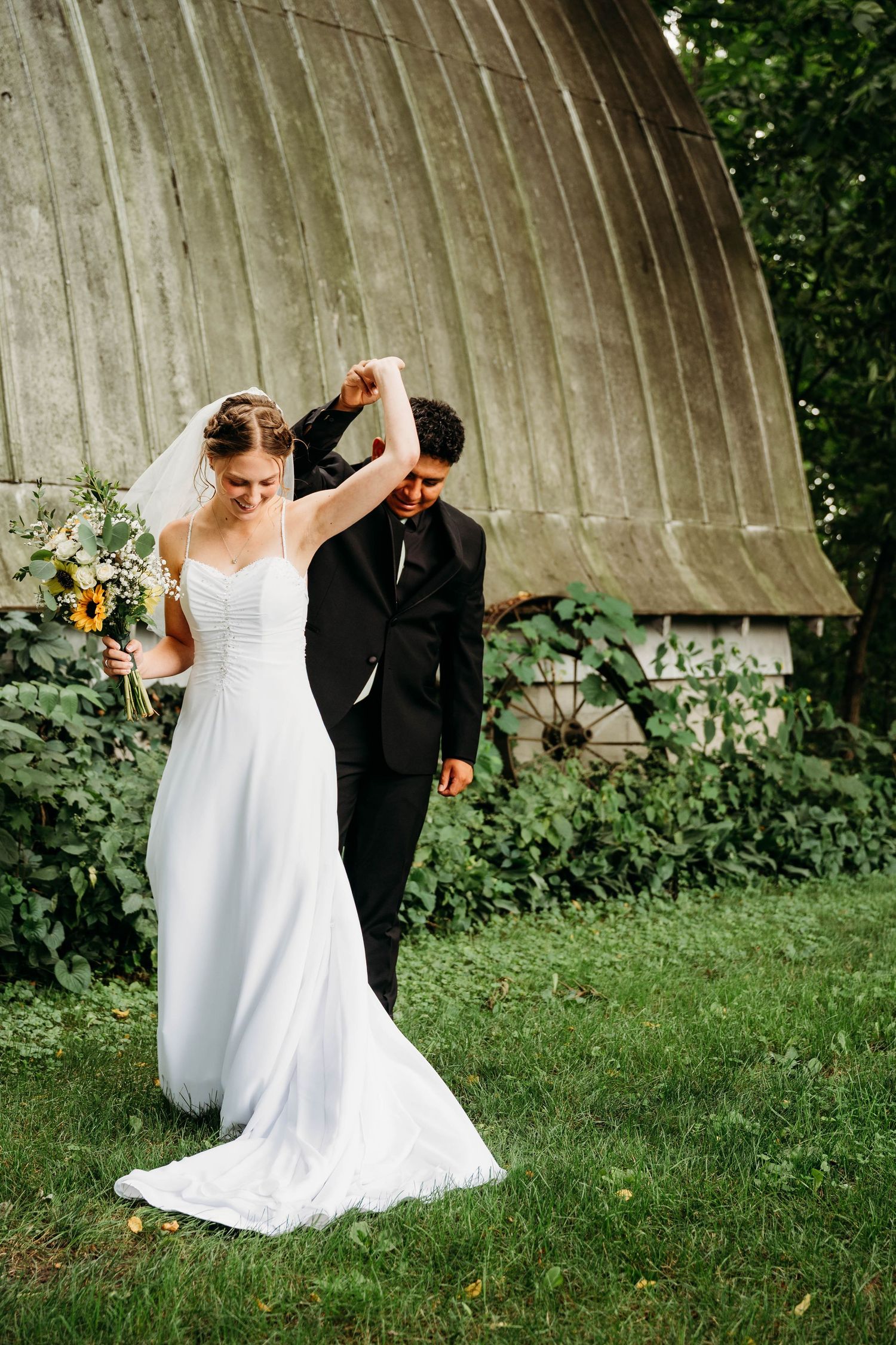 Bride and groom dancing outdoors near a rustic barn, bride in white gown holding sunflower bouquet.