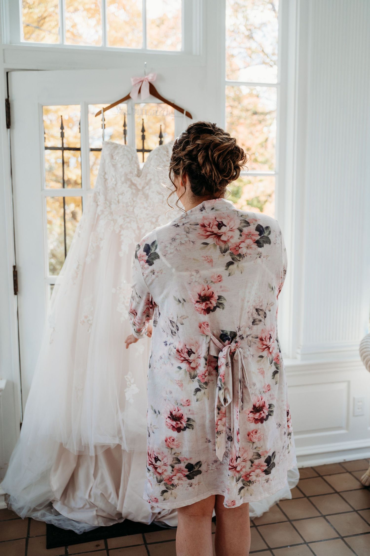 Someone in a floral robe gazes at an elegant white lace wedding dress hanging in sunlit window.
